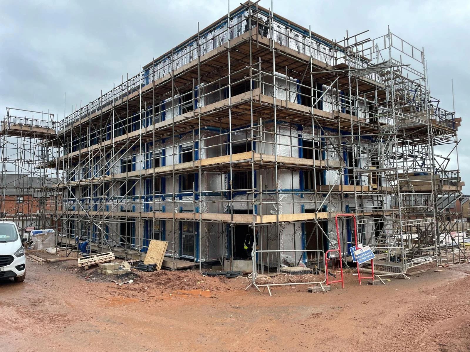 A building under construction with multiple levels of scaffolding surrounding it. The exterior is covered with insulation material, and construction equipment, including barriers, is visible at the site.