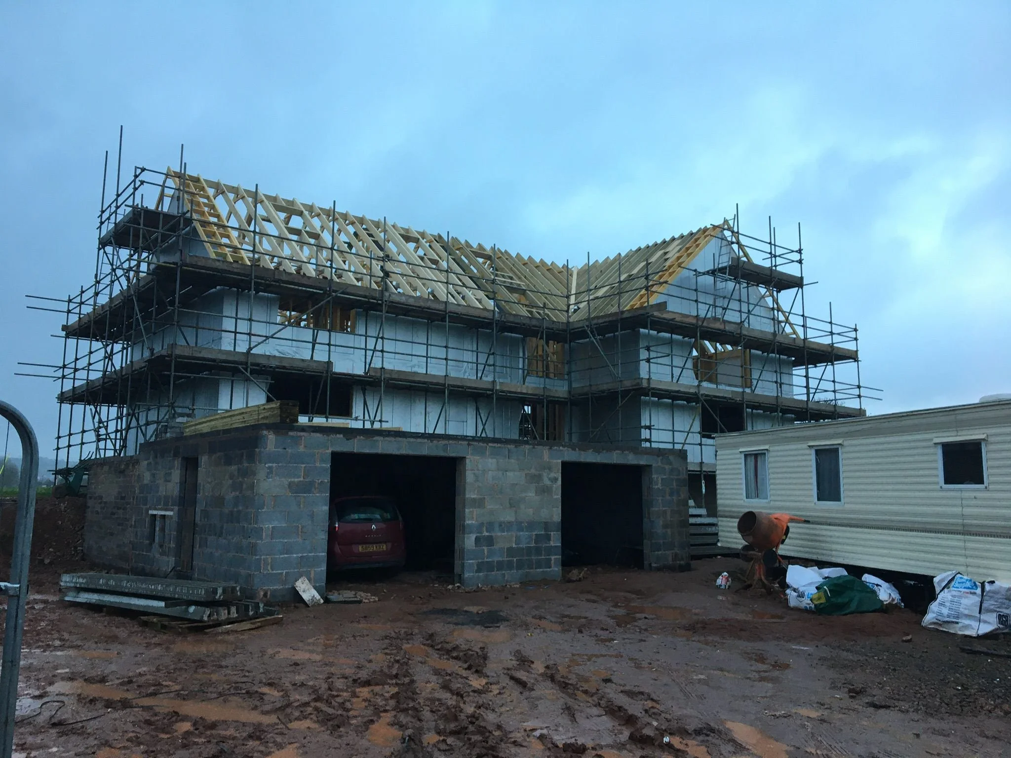 A house under construction with scaffolding around it, showing the wooden roof framework and part of the concrete foundation, with construction materials and equipment nearby.