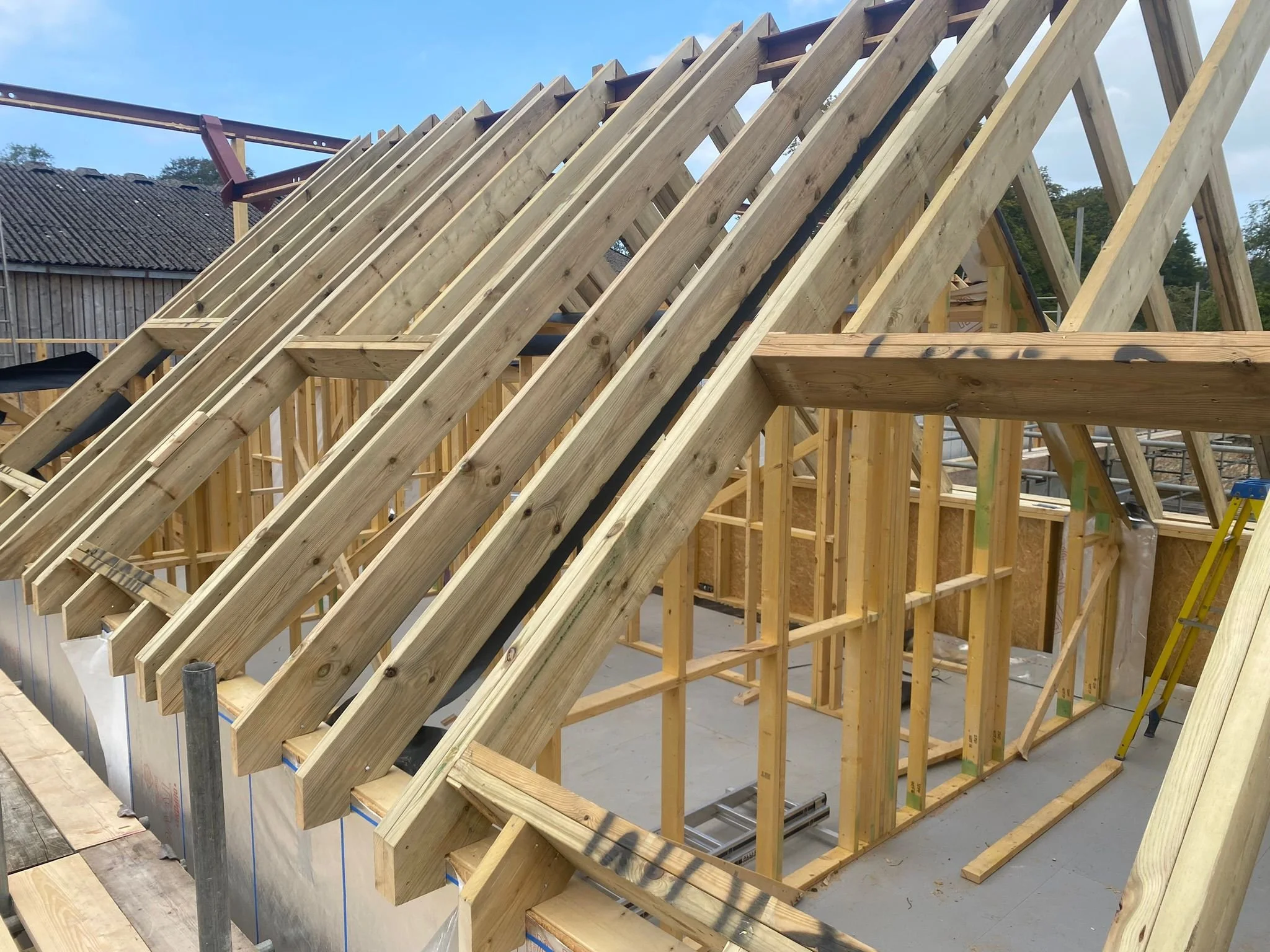 Construction site of a building with wooden framing and roof trusses under construction, with a clear sky in the background.
