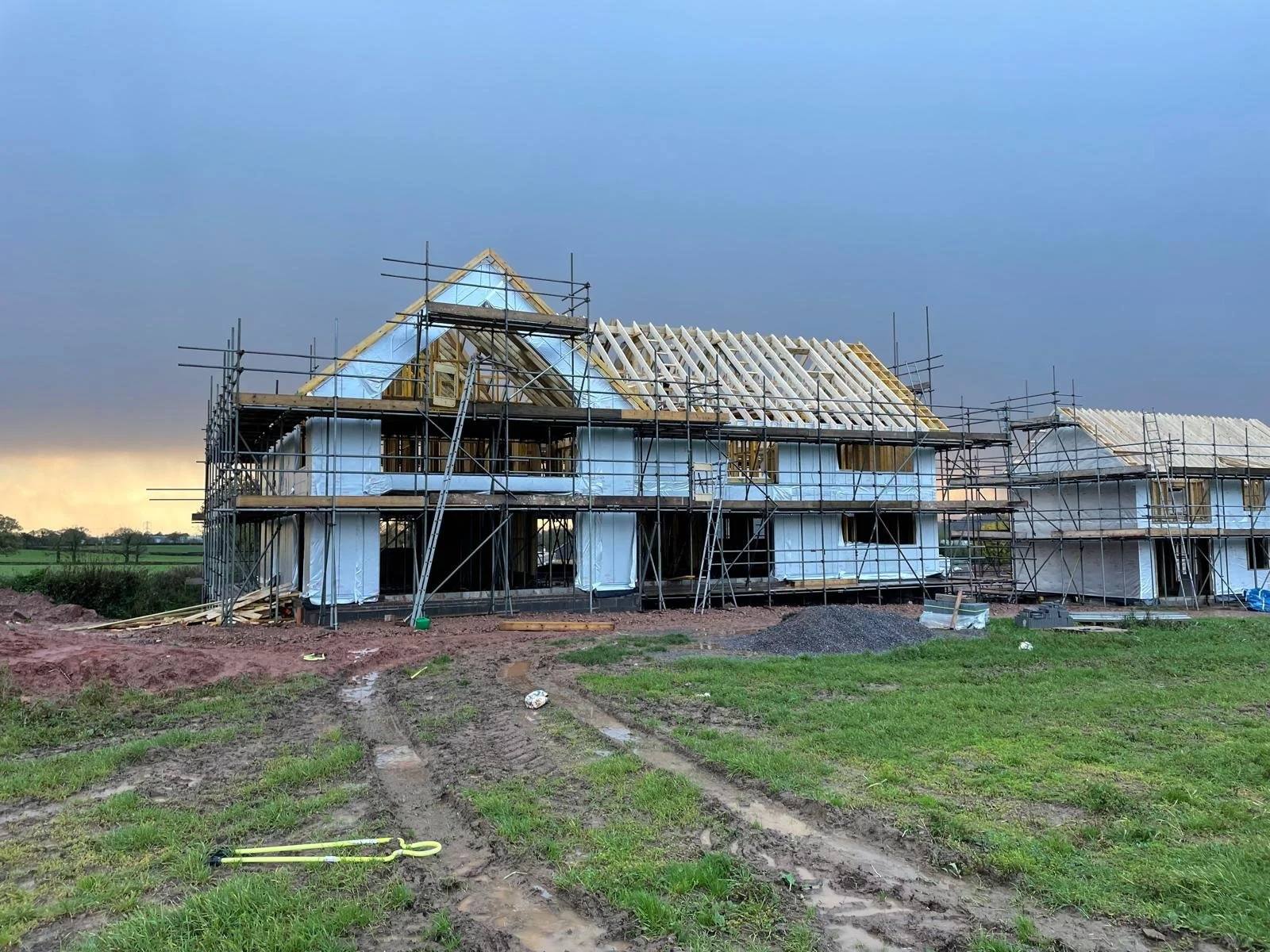 Under construction house with scaffolding, exposed wooden framing, and partially finished roof, set on a grassy area with muddy tracks in the foreground and a cloudy sky in the background.