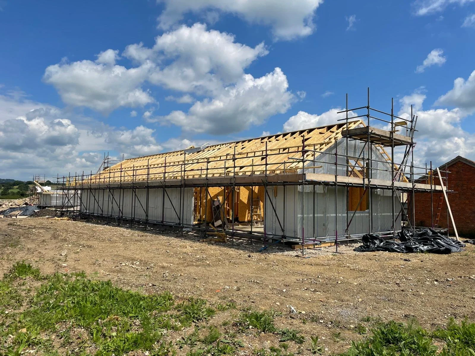 A building under construction with scaffolding around it, a wooden roof structure, and newly framed walls. The sky is partly cloudy with green landscape in the background.