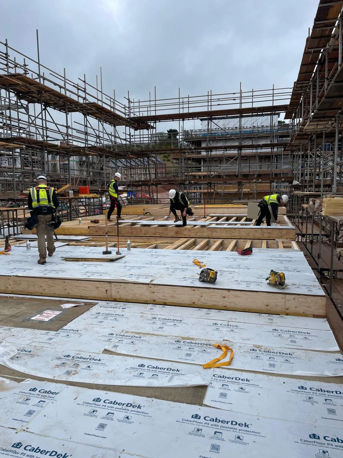 Construction site with workers installing flooring, surrounded by scaffolding and building framework, under an overcast sky.