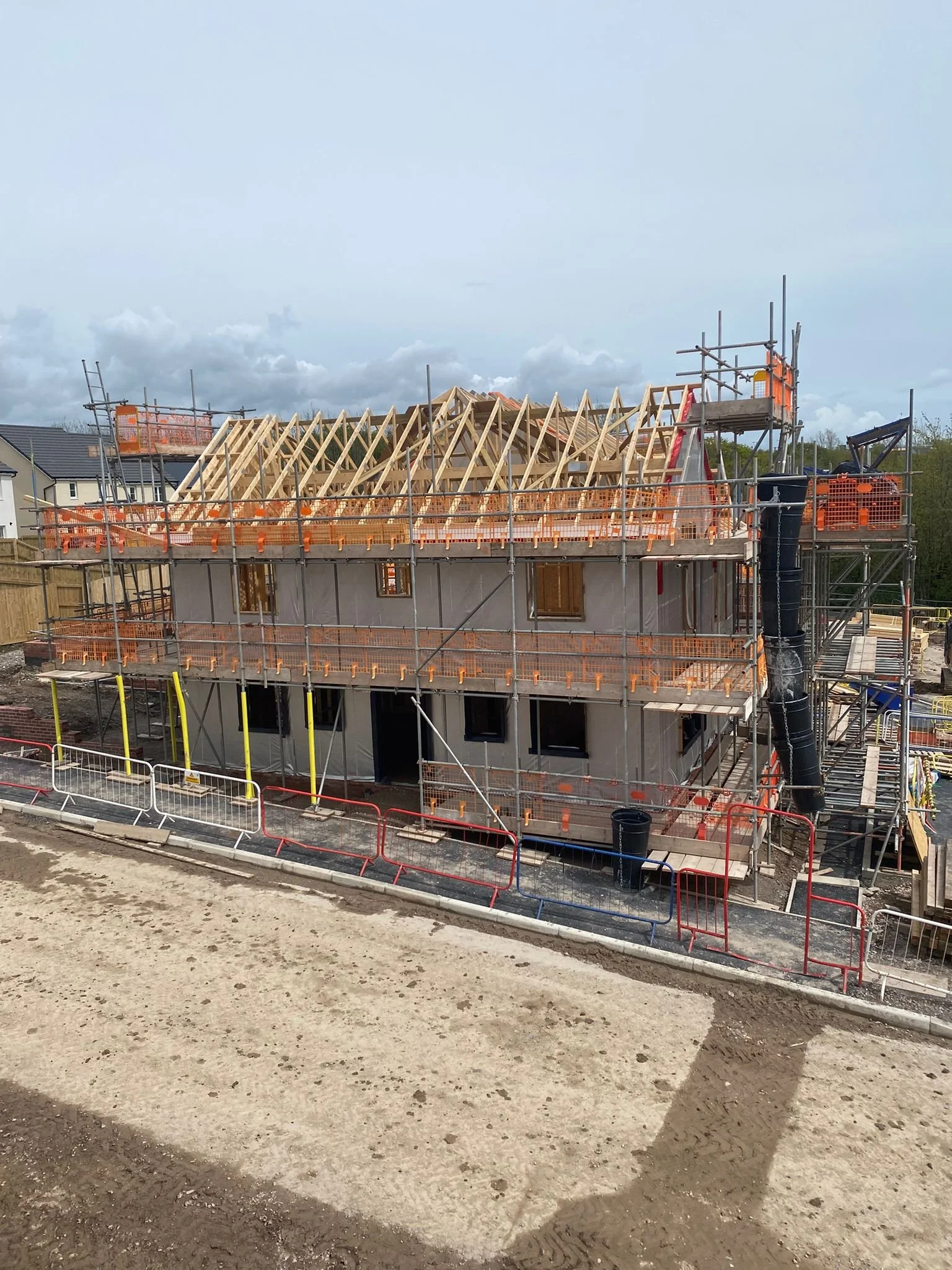 A two-story house under construction with visible framing and roof trusses, surrounded by scaffolding and safety barriers.