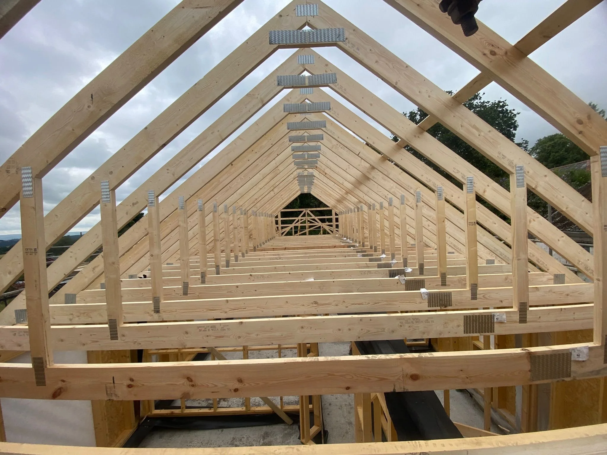 View of a house roof framing under construction with wooden beams and trusses, set against a cloudy sky.