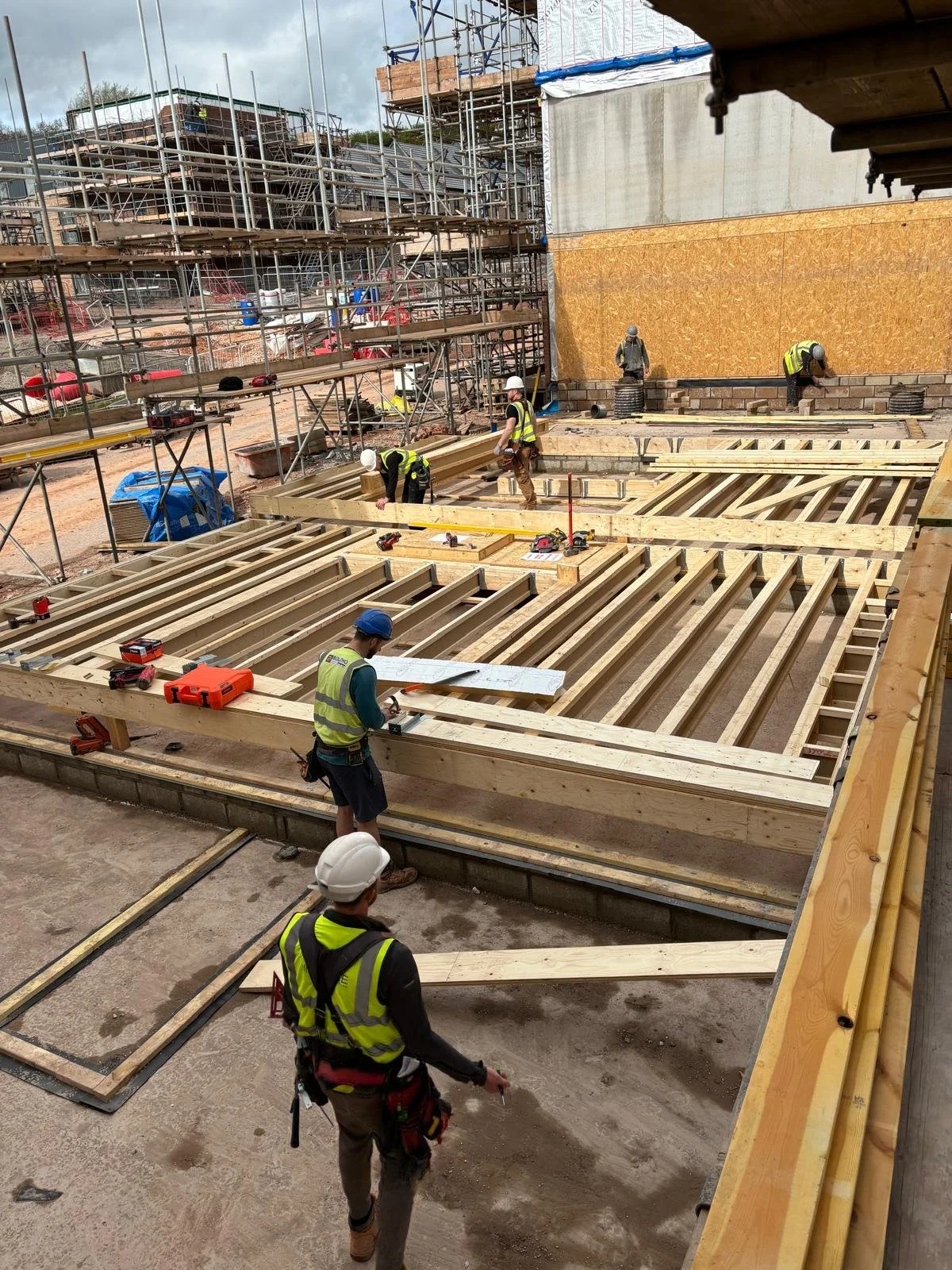 Construction workers building a wooden floor frame on a construction site, with scaffolding and partially built structures in the background.