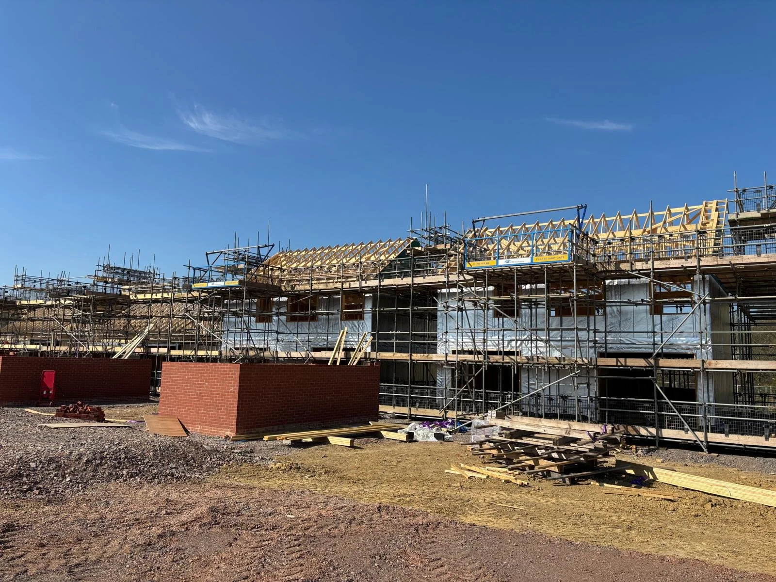 Building under construction with scaffolding, wooden roof framework, and brick walls, under a clear blue sky.