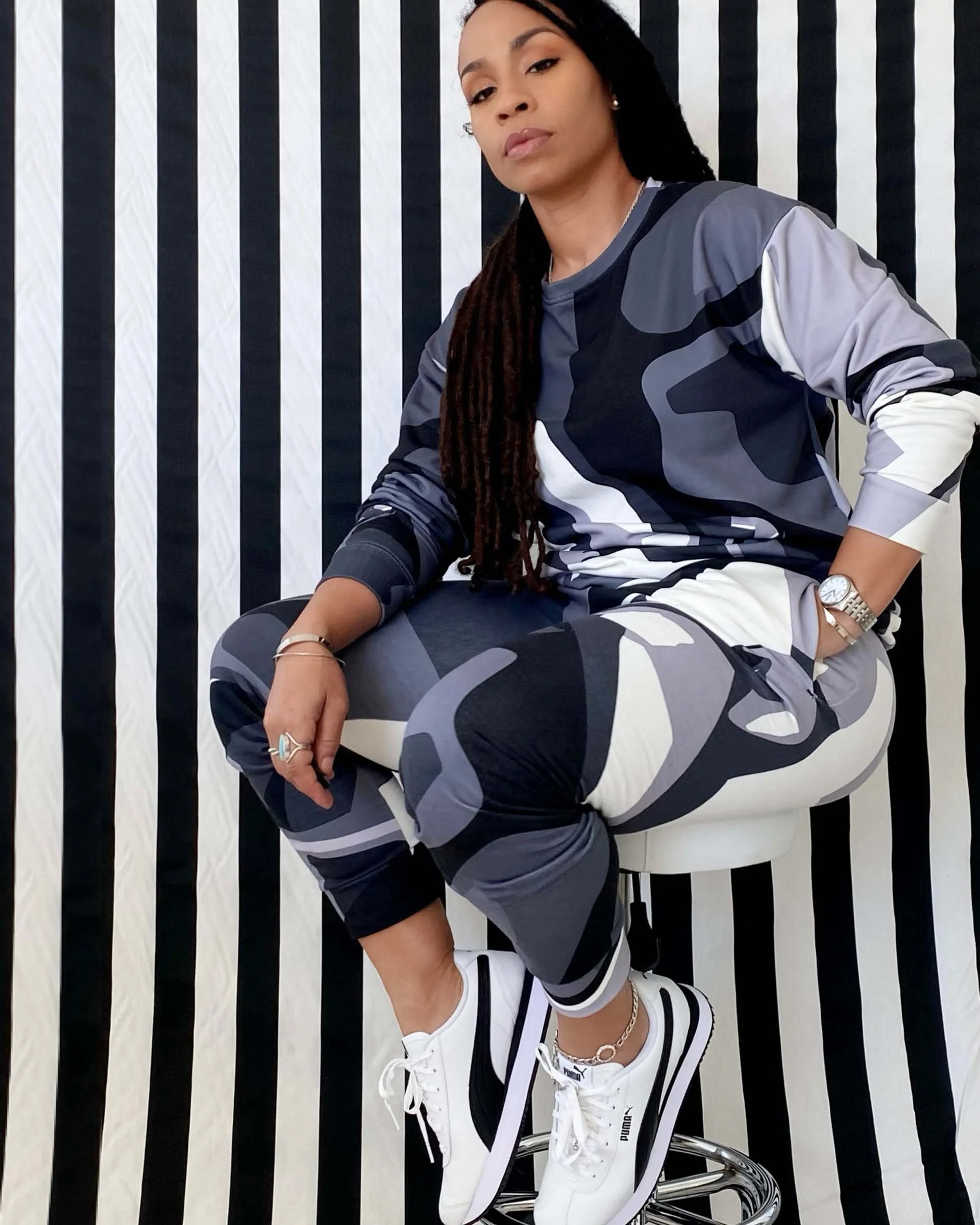 A woman with long dreadlocks, wearing a black, gray, and white camouflage hoodie and matching shorts, sitting on a metallic chair in front of a black and white striped background. She is wearing white sneakers, a watch, rings, and has a neutral expression.