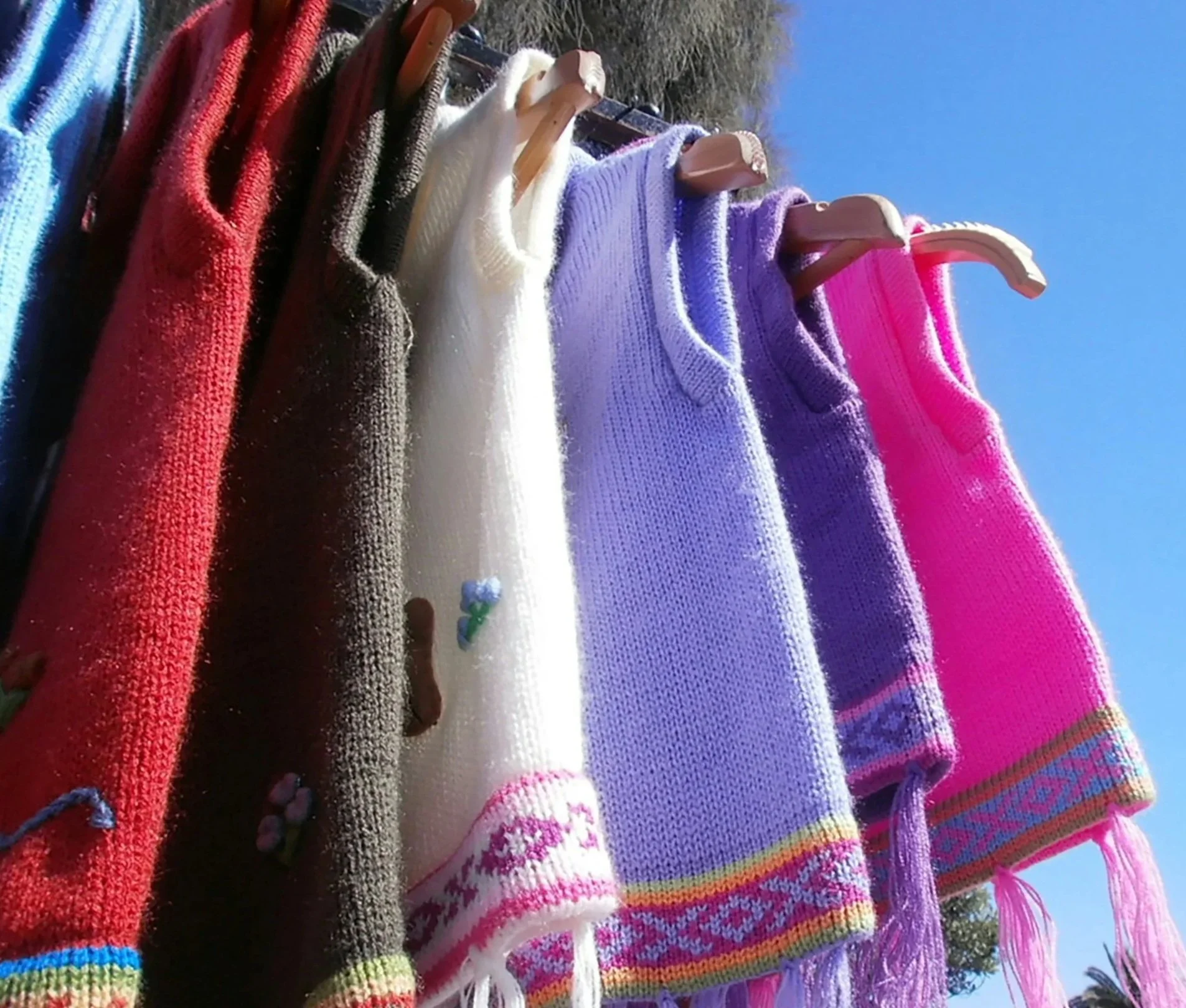 Colorful knit sweaters hanging on wooden hangers outdoors against a clear blue sky.