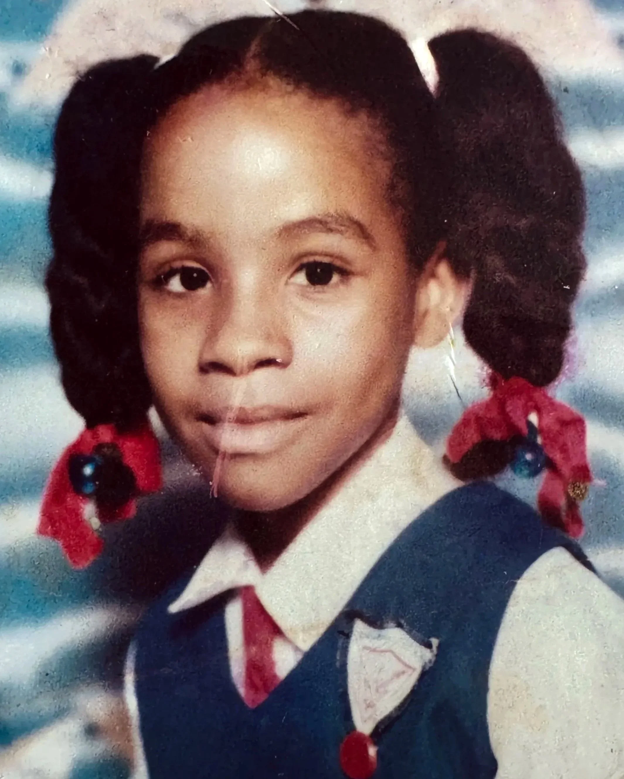 School portrait of Kimlayn as a child in Jamaica, wearing her Lannaman’s Preparatory School uniform with red ribbons in her hair.