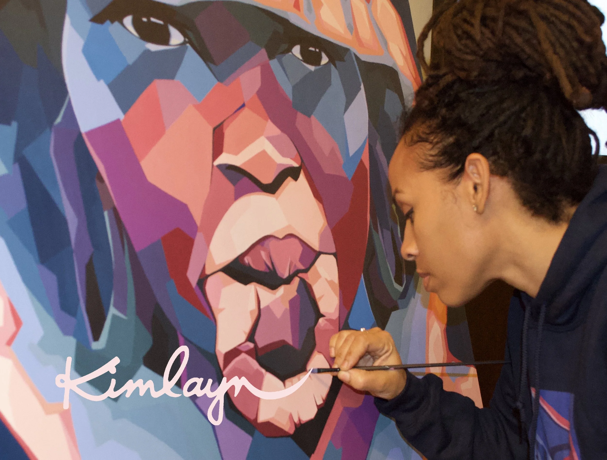 A woman with dark hair styled in dreadlocks is seen in profile painting a colorful geometric portrait of a woman with dark hair and brown eyes on a pink background. The woman is holding a paintbrush and is focused on her artwork.
