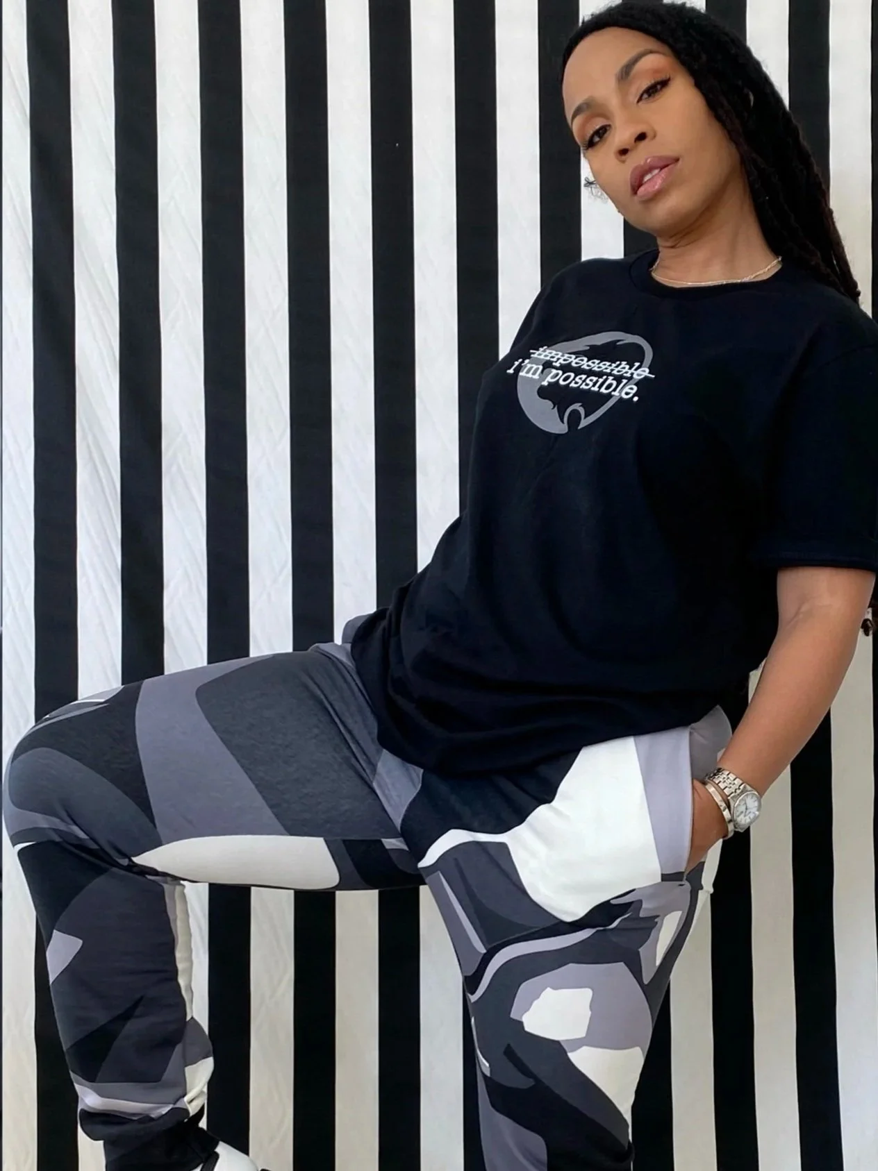 A woman wearing a black T-shirt with a logo and slogan, camo-patterned sweatpants, a watch, and jewelry, seated against a black and white striped background.