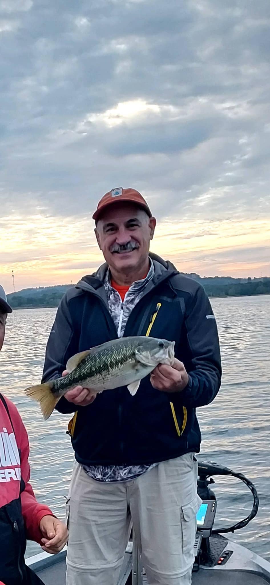 Man smiling and holding a largemouth bass fish on a boat during sunset.
