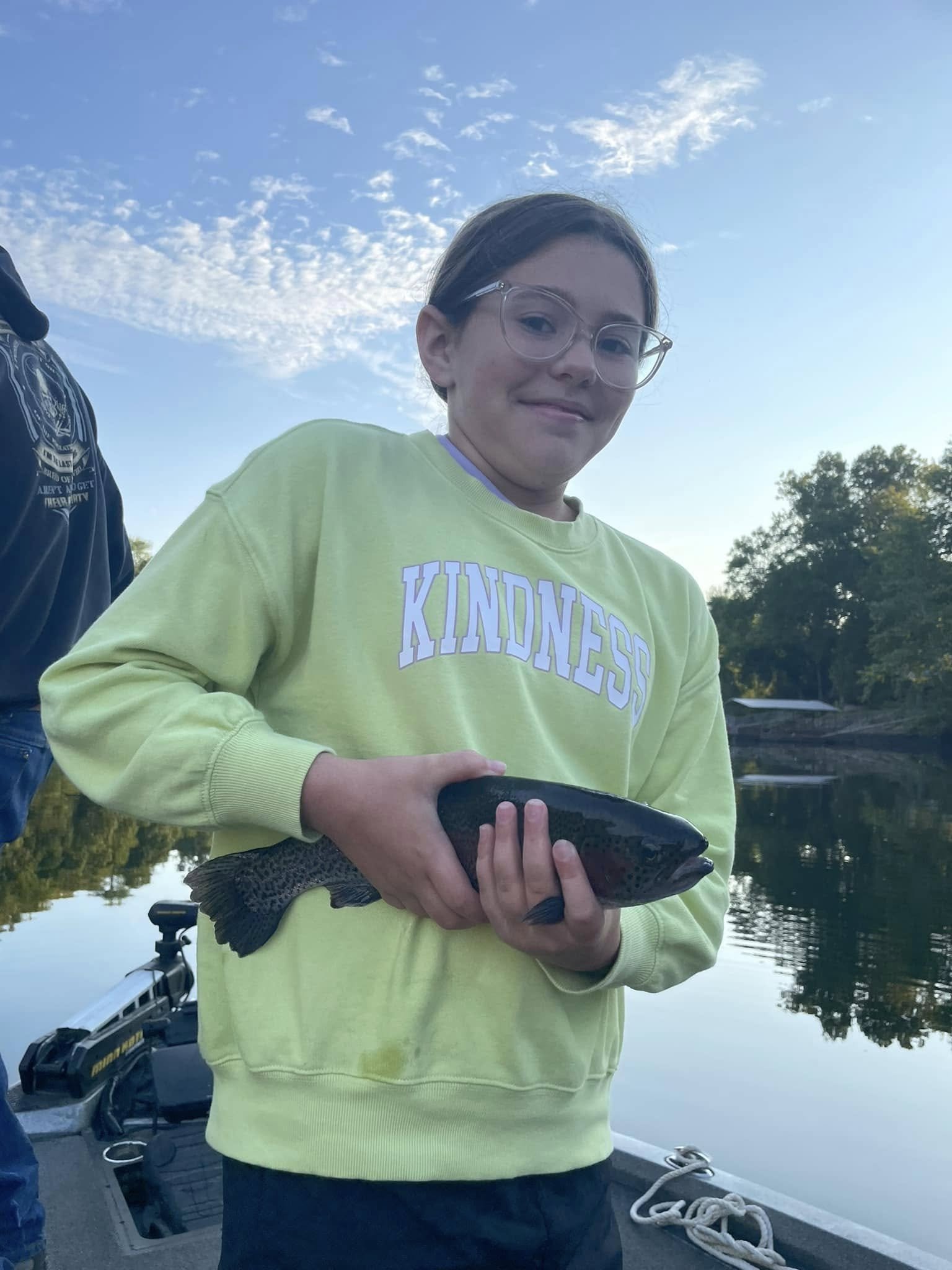 A young girl in a yellow hoodie with the word 'KINDNESS' printed on it, holding a large fish while standing on a boat near a calm lake during daytime with trees in the background.