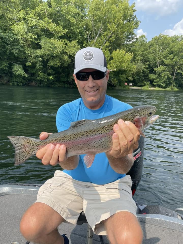 A man wearing sunglasses and a blue shirt holding a rainbow trout on a boat in a river with green trees in the background.