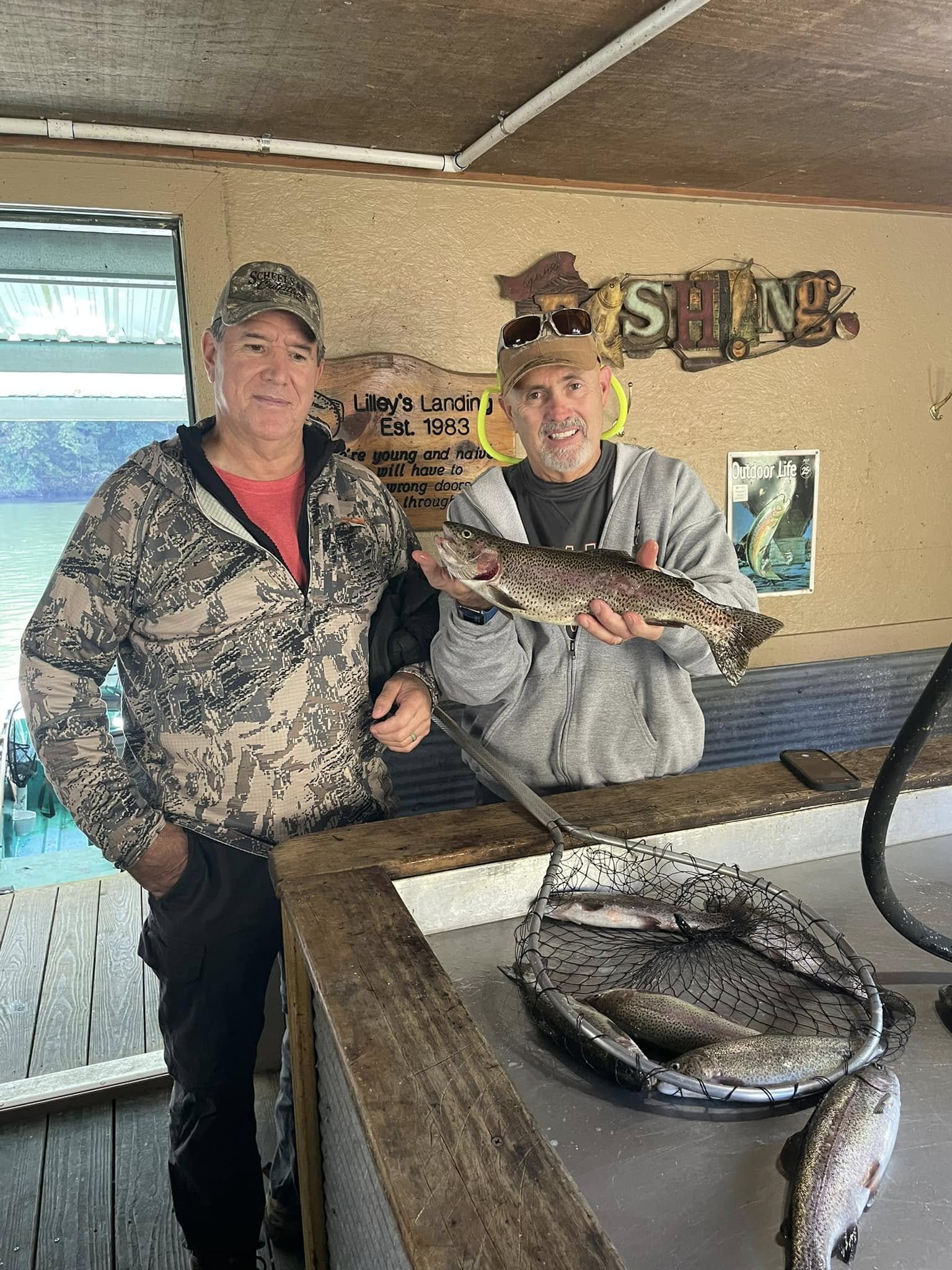 Two men posing indoors with a caught fish, one holding a large rainbow trout, after a fishing trip at a lake.