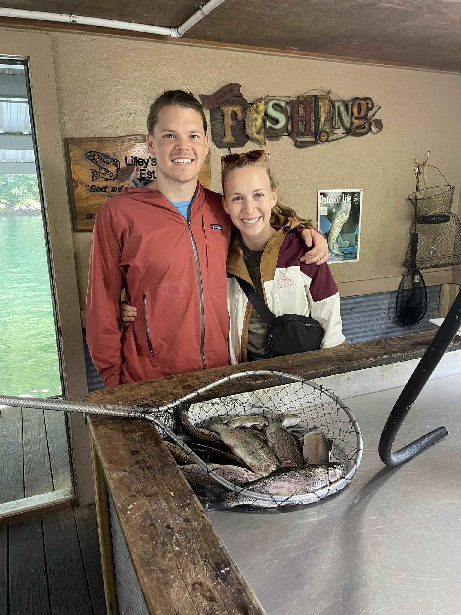 A smiling man and woman standing behind a counter with a basket of freshly caught fish, inside a rustic fishing lodge or cabin.