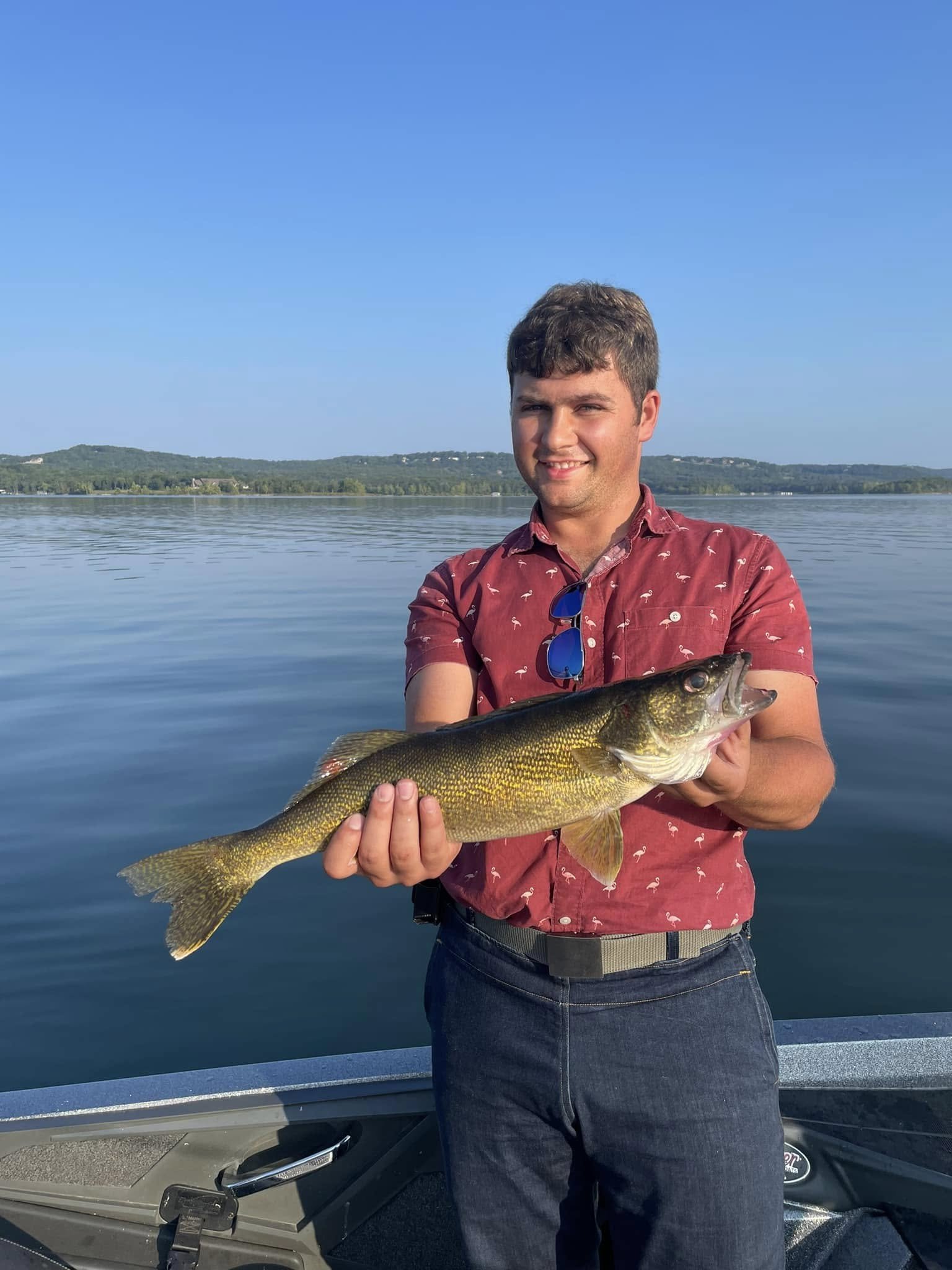 A young man in a red shirt with flamingo patterns holding a large fish on a boat, with a calm lake and distant hills in the background under a clear blue sky.