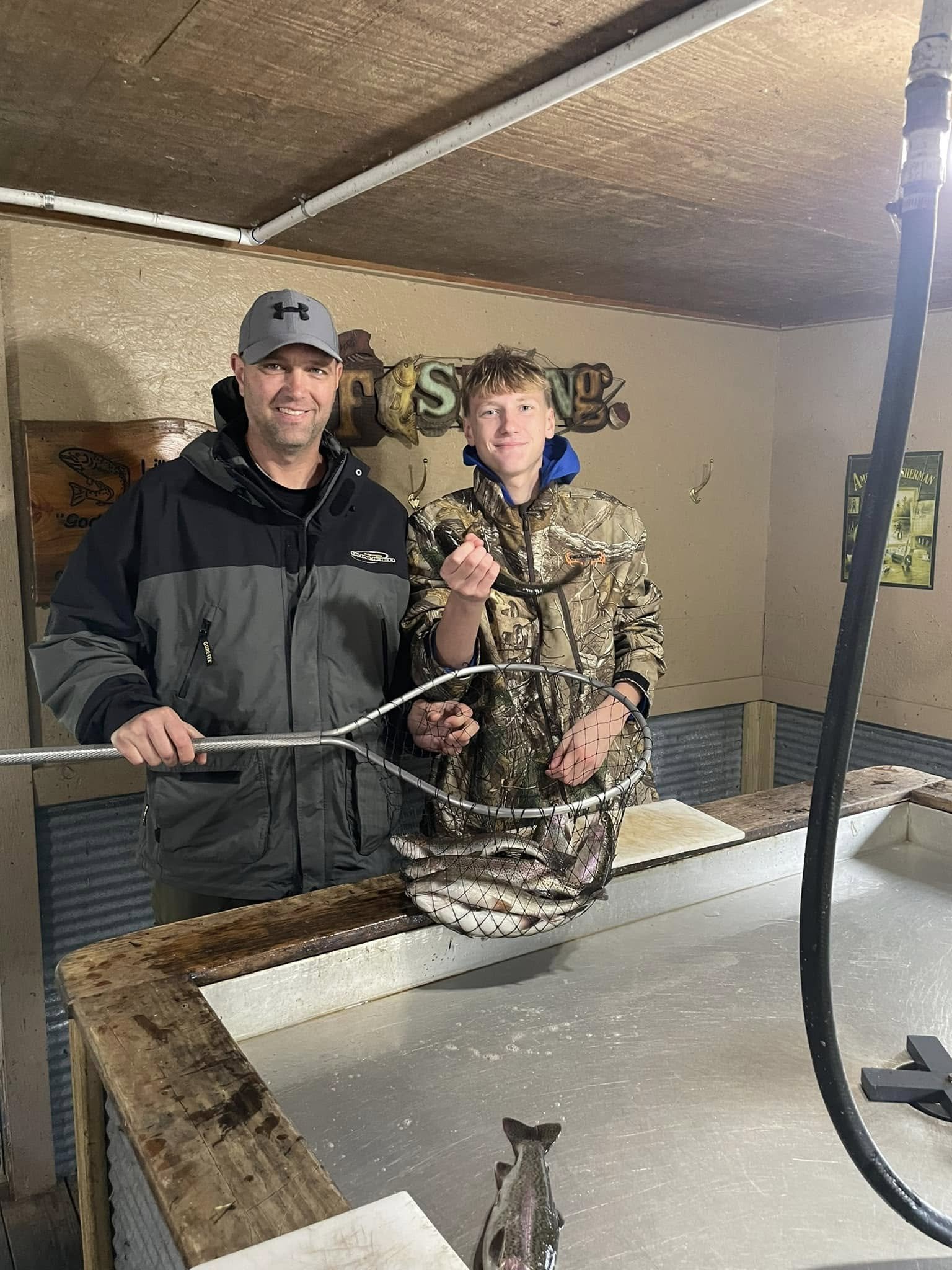 Two males standing behind a fish cleaning station, holding a fishing net with multiple fish inside. One is an adult wearing a gray and black jacket and a gray cap, the other is a young boy in camouflage clothing. The younger boy is holding a fish in