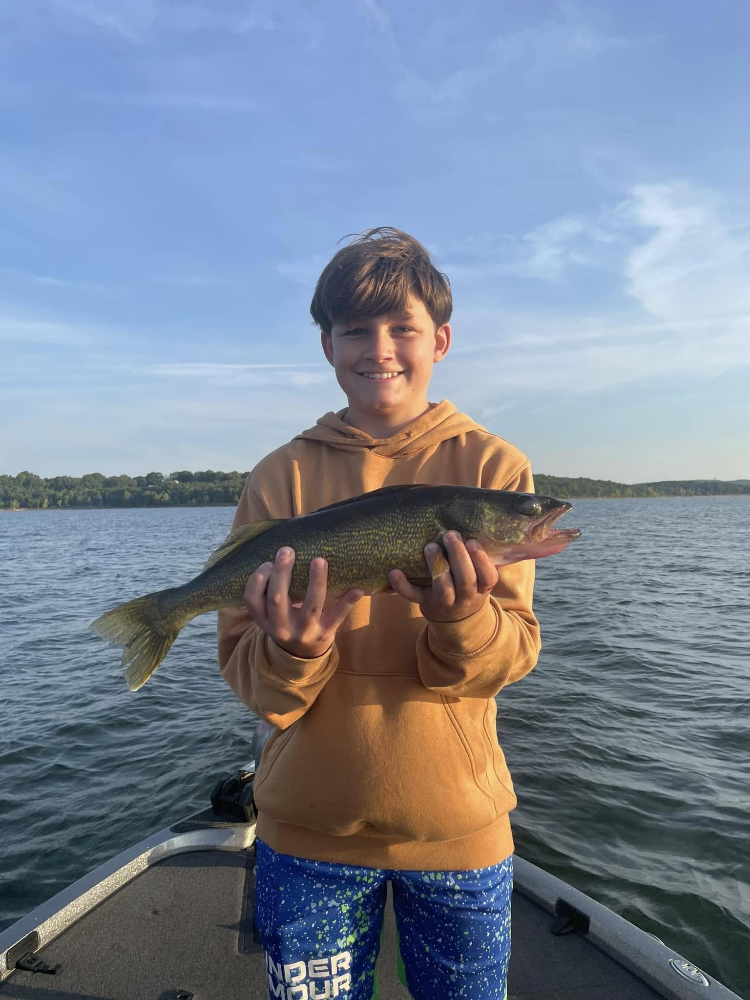 A young boy in an orange hoodie and blue patterned shorts standing on a boat, holding a large fish over the water with a cloudy sky in the background.