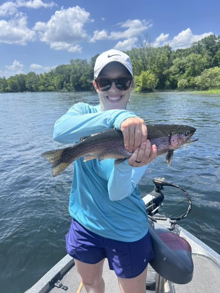 A woman wearing a white Nike cap, sunglasses, a blue long-sleeve shirt, and purple shorts standing in a boat on a lake holding a large rainbow trout fish with a natural background of trees and blue sky.