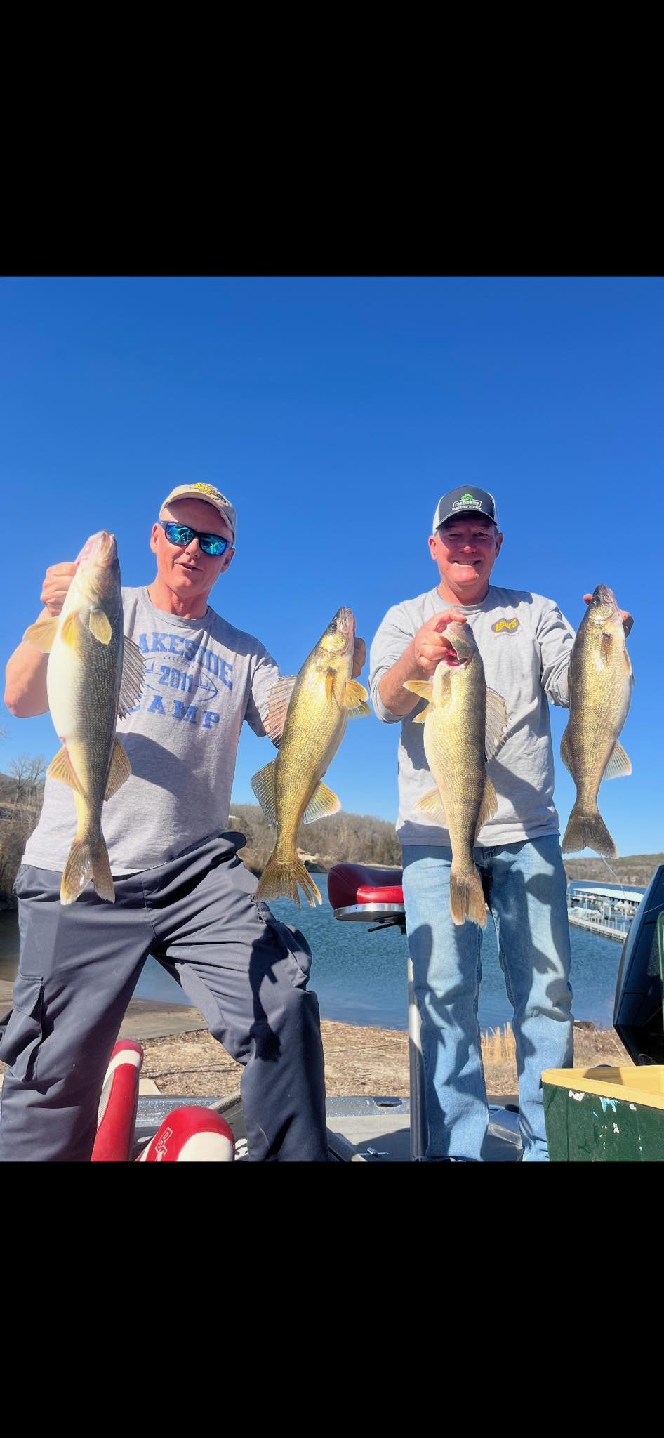 Two men standing outdoors holding up four large fish they've caught, with a lake and trees in the background under a clear blue sky.
