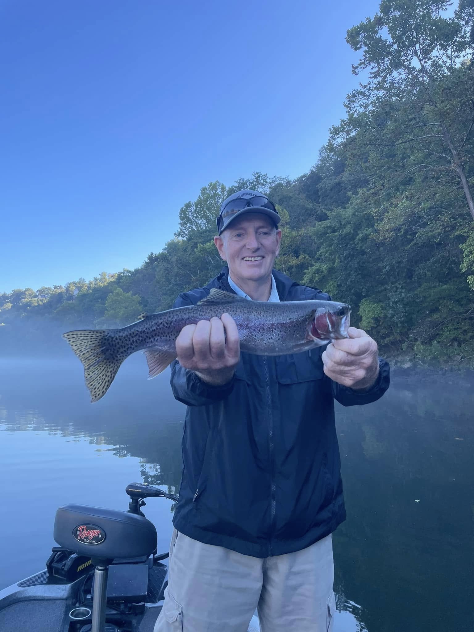 Man holding a large fish he caught while standing on a boat on a calm river with trees in the background under a clear blue sky.