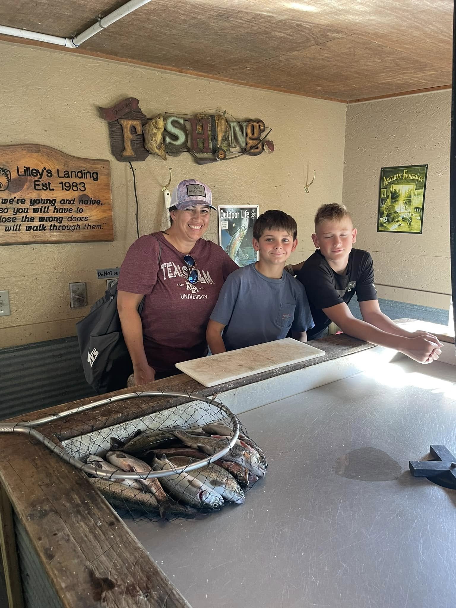 A woman and two boys smiling at a fish cleaning station with a basket of fish, wooden signs, and fishing-themed decorations in the background.