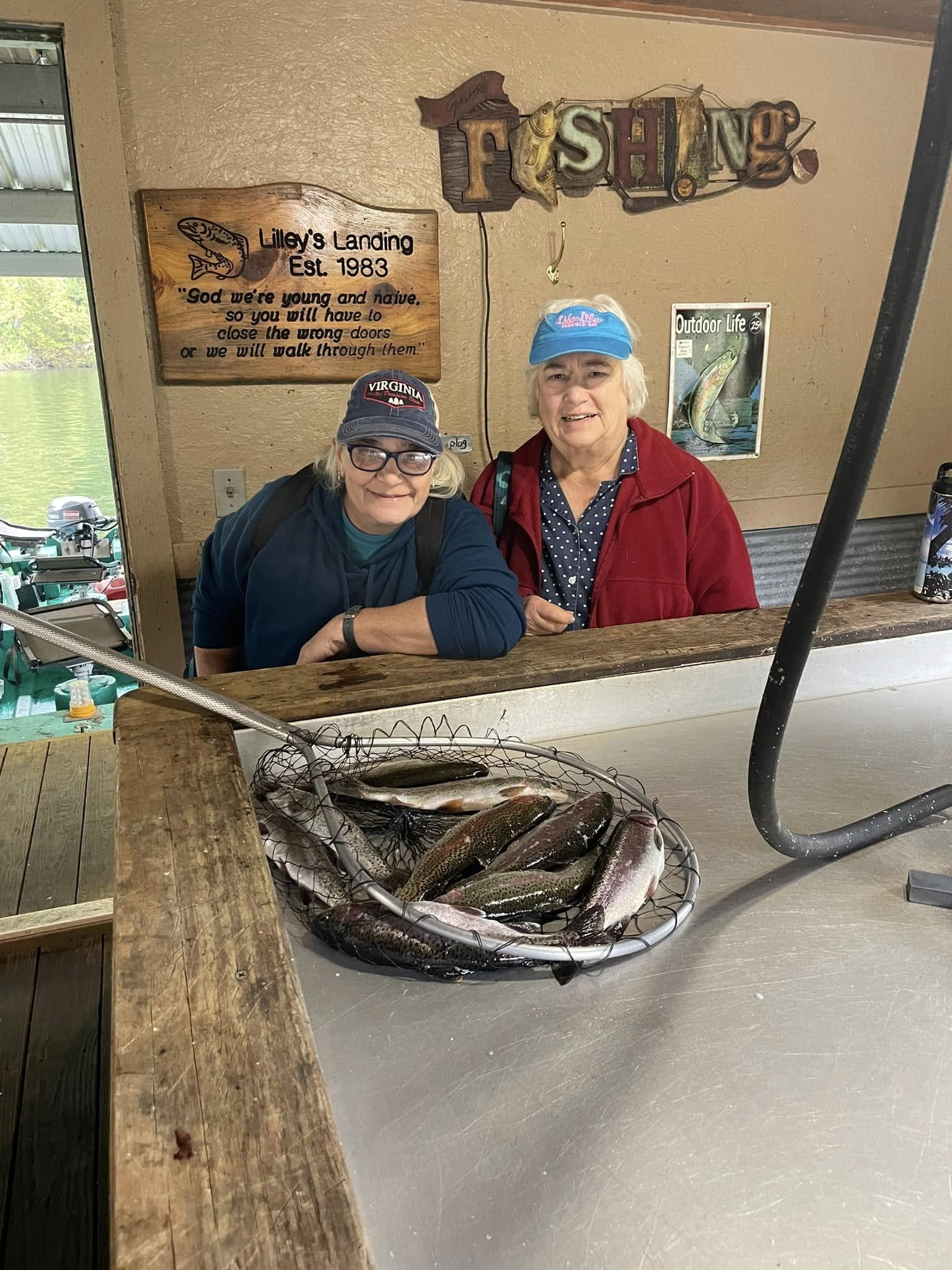 Two women at a fishing lodge behind a counter with a large basket of freshly caught fish, smiling at the camera.