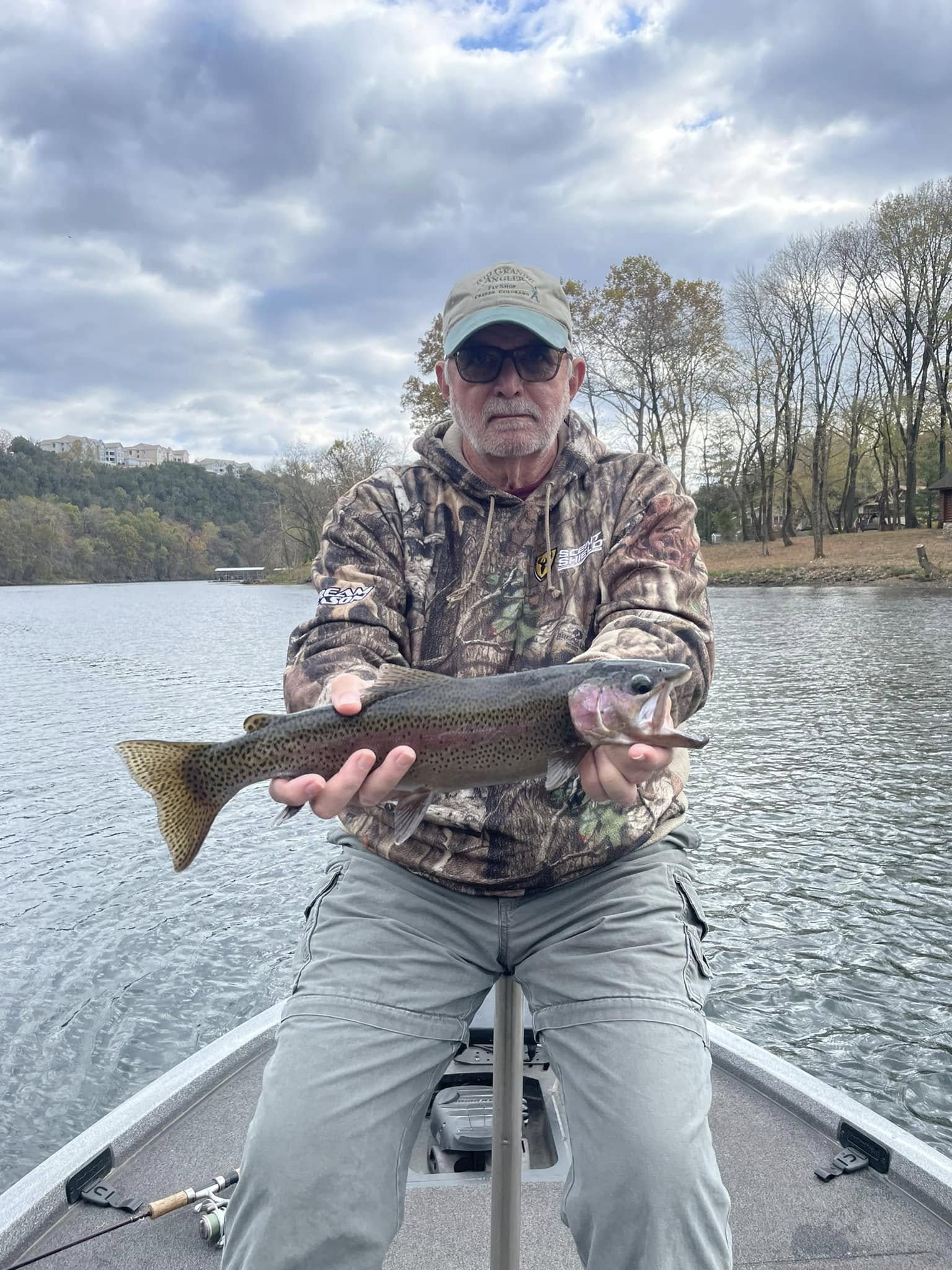 Man in camouflage hoodie, gray pants, and sunglasses holding a large rainbow trout on a boat on a river with trees and cloudy sky in the background.