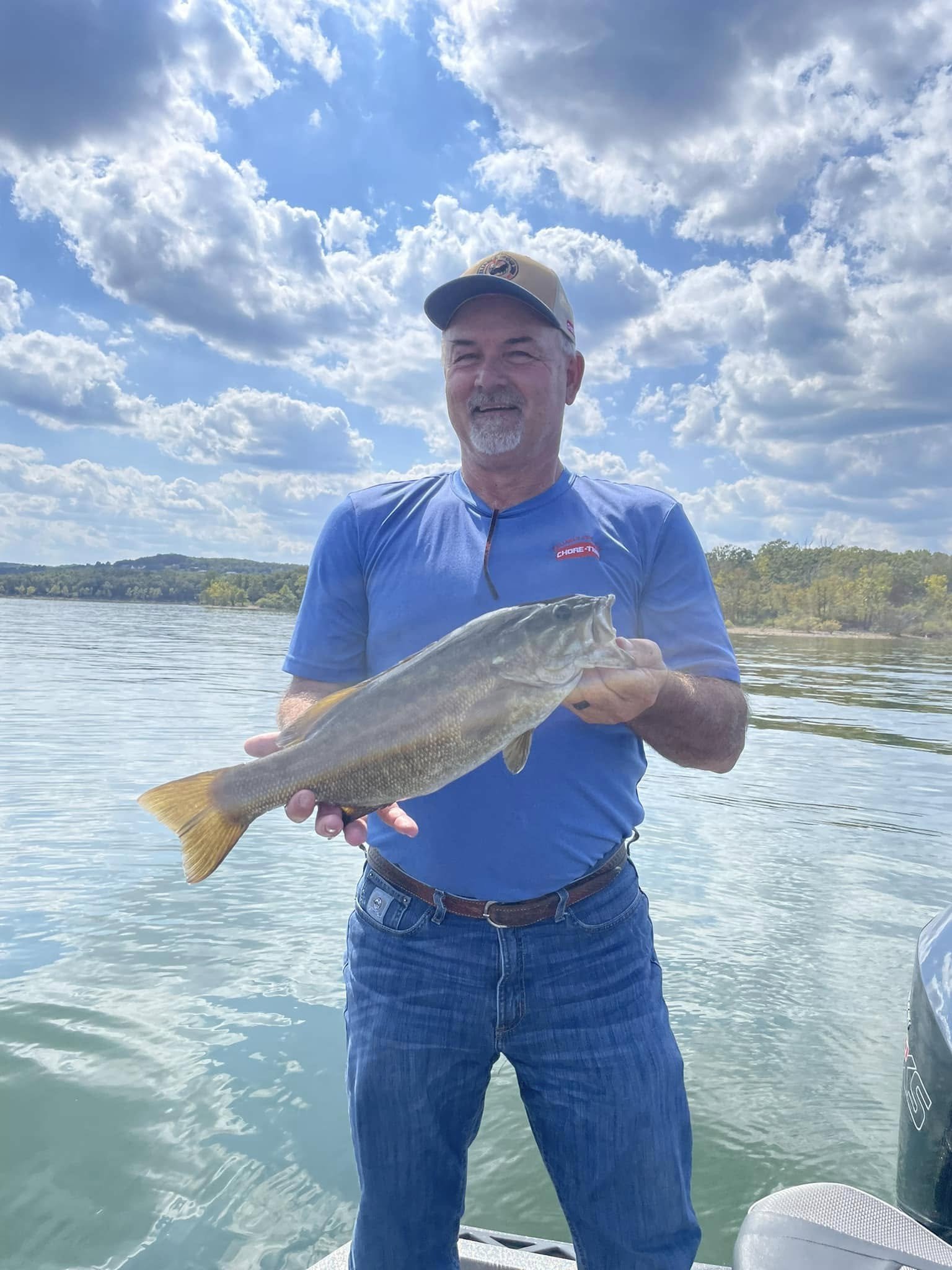 A man wearing a blue t-shirt and a cap is standing on a boat, holding a large fish he caught, with a lake and a partly cloudy sky in the background.