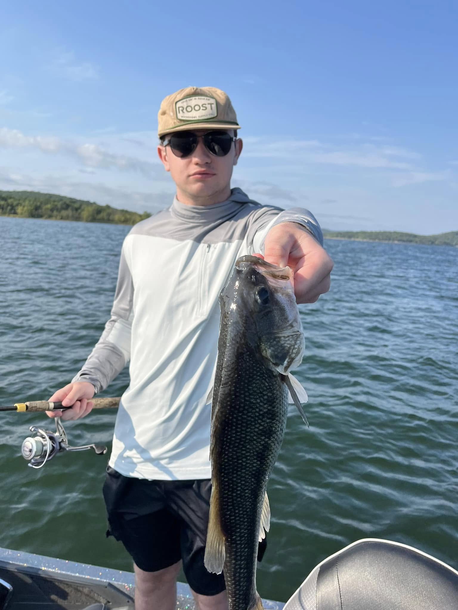 A man wearing sunglasses, a tan cap, and a gray and white long-sleeve shirt holding a large fish on a boat in a lake, with water and hills in the background.