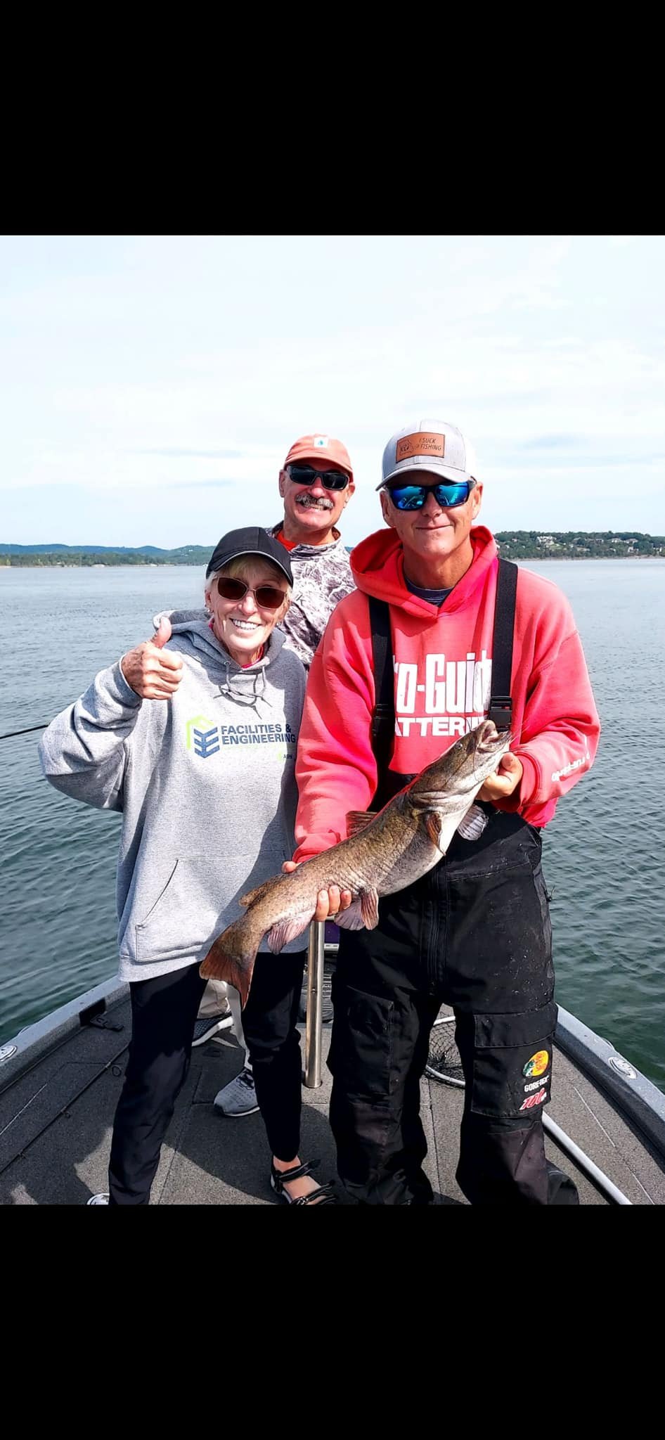 Three people on a boat, two men and a woman, holding a large fish and posing for a photo. The man in the front is wearing a red hoodie and sunglasses, holding the fish. The woman and the other man are behind him, smiling and giving a thumbs-up, with