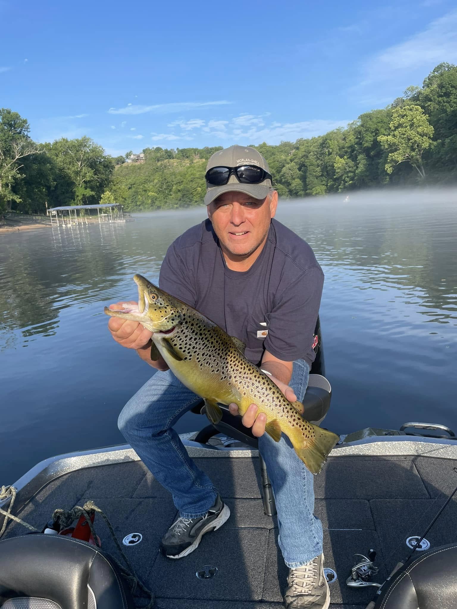 A man in a boat holding a large, freshly caught brown trout with a scenic lake and green trees in the background.