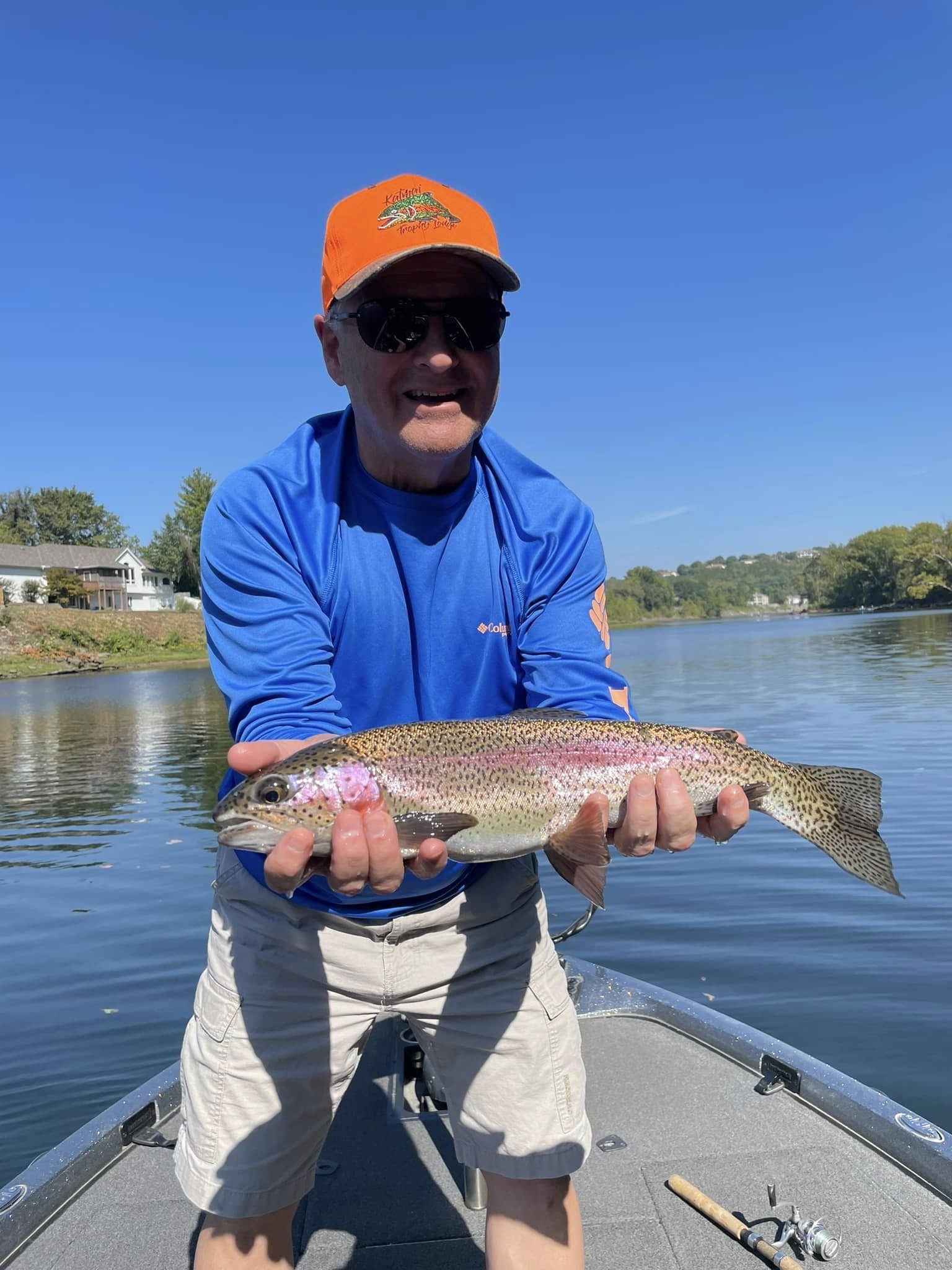 A man wearing sunglasses, a bright orange cap, a blue long sleeve shirt, and beige shorts is holding a large rainbow trout fish on a boat on a calm body of water on a sunny day.