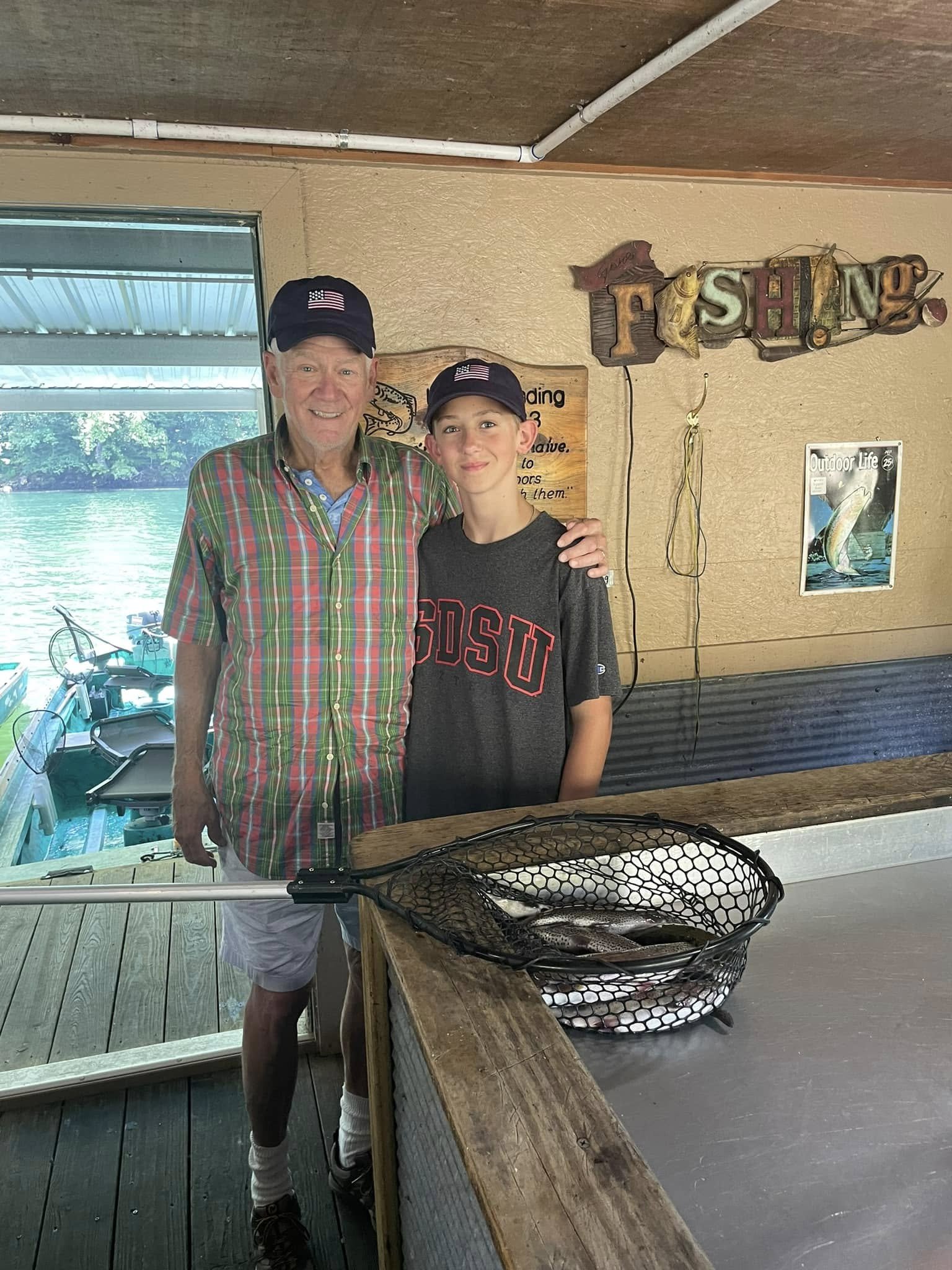 An elderly man and a teenage boy standing together inside a fishing lodge next to a basket of caught fish, with fishing-themed decor on the wall, including a sign that reads 'FISHING.' They are both wearing black caps with American flags and smiling