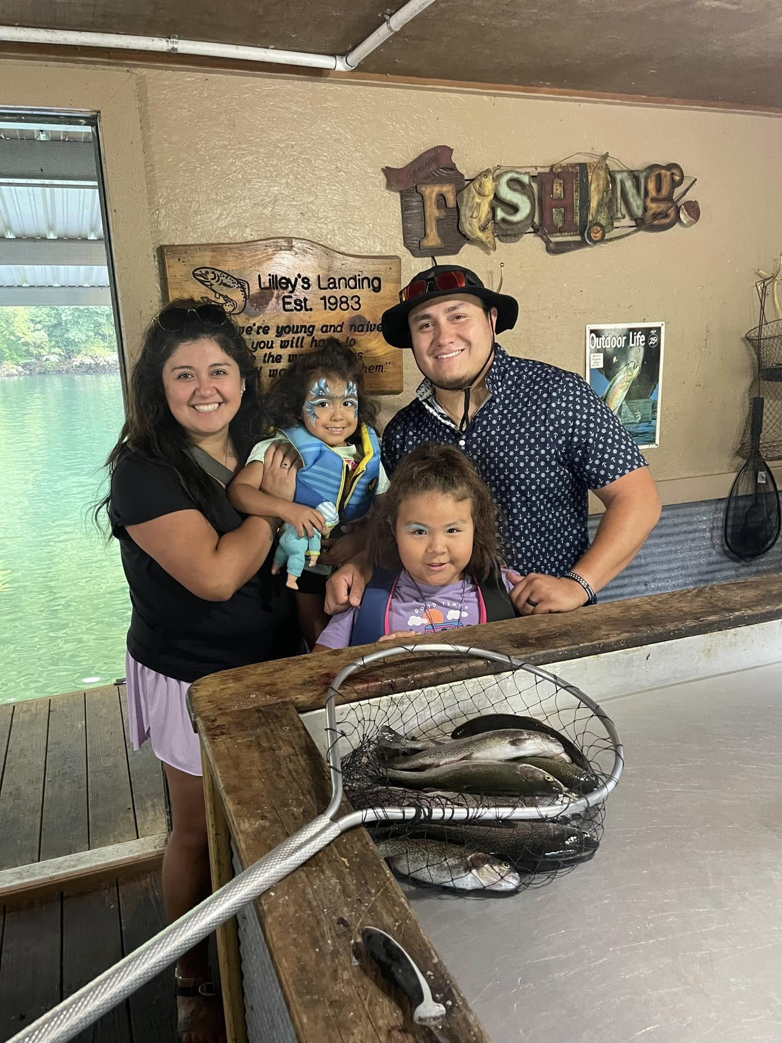 A family of four poses together inside at a fishing-themed location. The woman holds a young girl with face paint, and an older girl stands in front. A man in a hat smiles next to them. Fish are displayed on a net on the table in front of them.