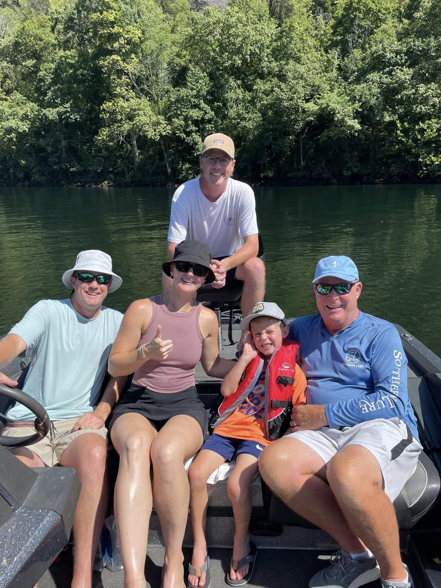 Family enjoying a boat ride on a river with lush green trees in the background. The group includes two men, two women, and a young boy, all smiling and wearing casual summer clothing and hats.