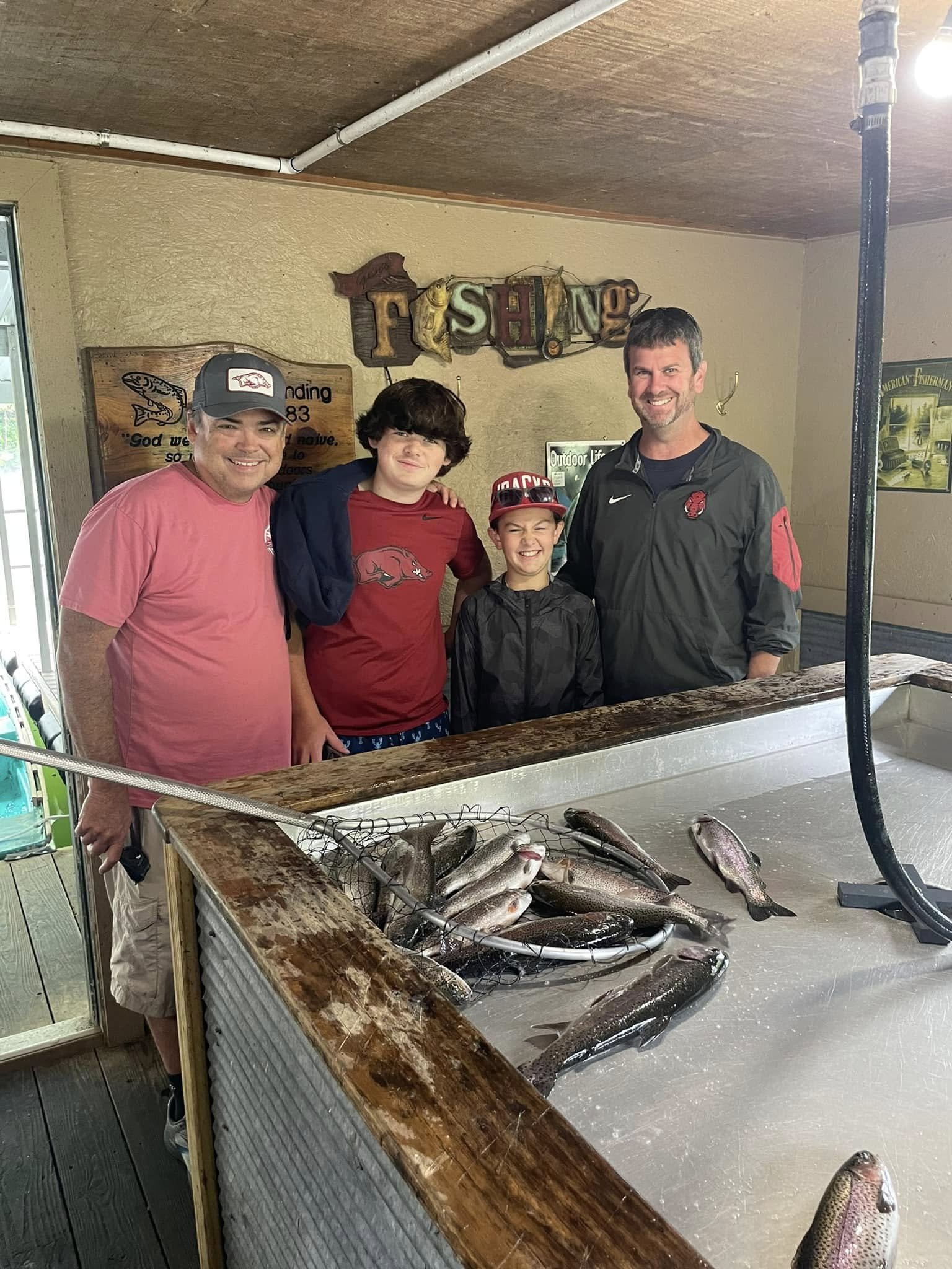 Four people standing behind a fishing display with several caught fish, in an indoor shop with a sign above that says 'FISHING' and fishing-related decor.