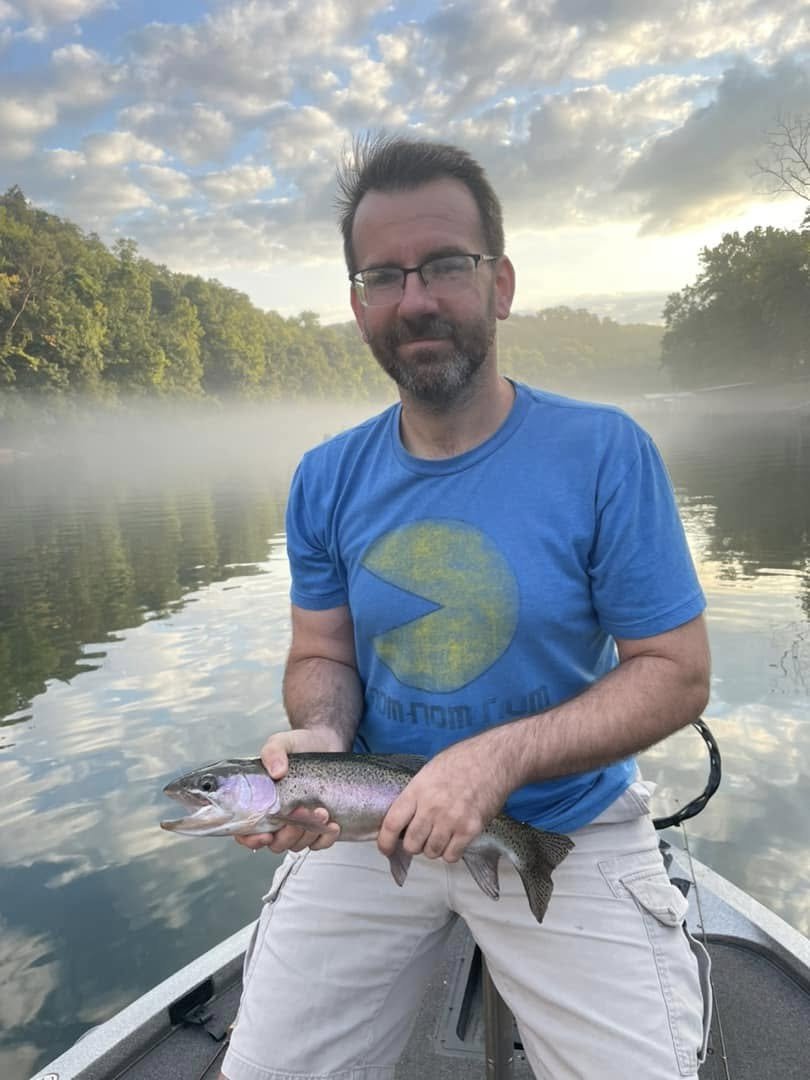 A man with glasses and a beard, wearing a blue Pac-Man shirt, holds a fish on a boat with a river and trees in the background during a cloudy morning or evening.