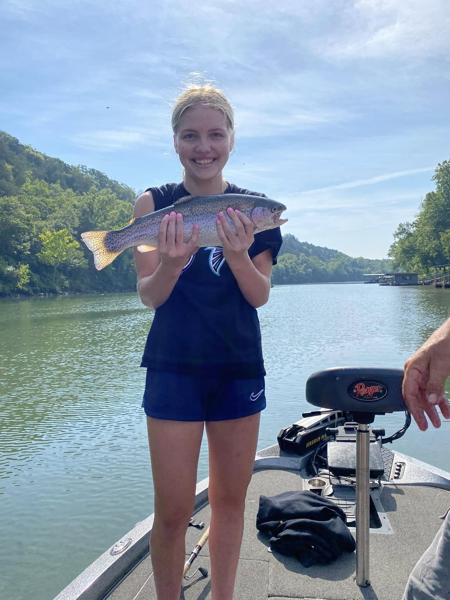 A girl on a boat holding a large rainbow trout she caught, with water and green trees in the background.