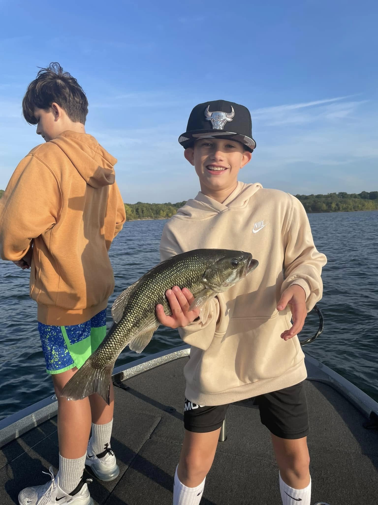 A young boy smiling and holding a large fish on a boat, with another boy standing nearby on a lake during daytime.