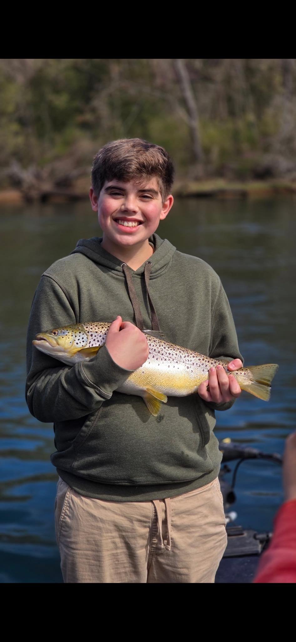 A smiling young boy holding a large rainbow trout near a body of water outdoors.