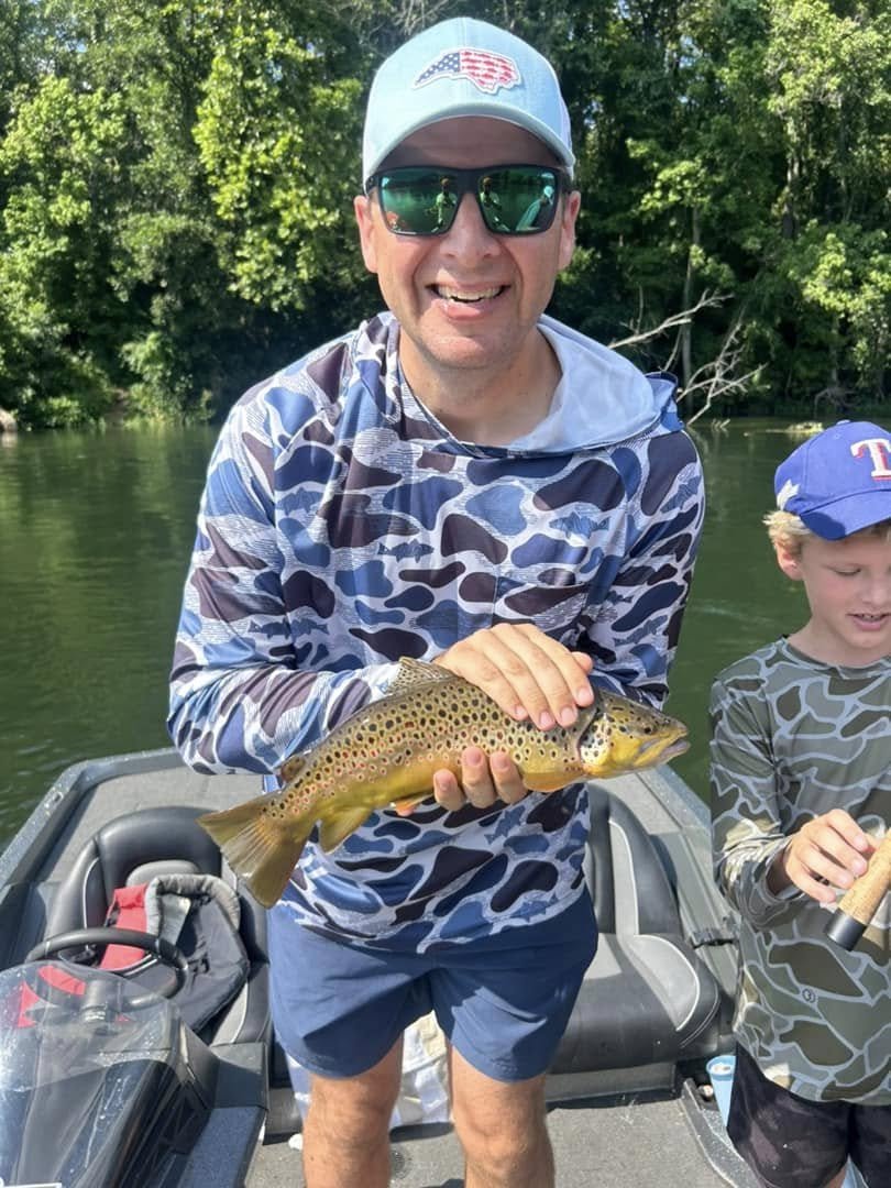 A man in sunglasses, a cap, and a camouflage-patterned shirt holding a fish on a boat, with a boy in a camouflage shirt and a blue baseball cap beside him, near a green forest background.