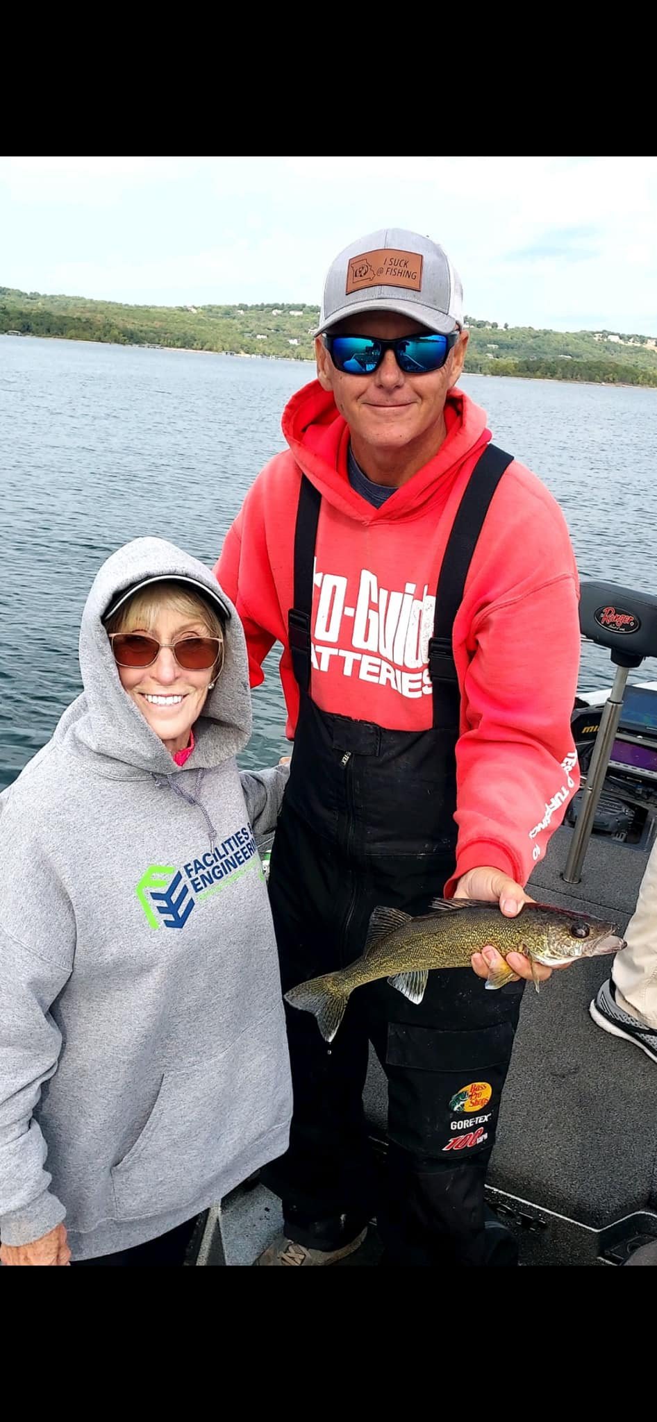A man and woman on a boat holding a fish, with water and hills in the background. The man is wearing a red hoodie, sunglasses, and a hat, and the woman is wearing sunglasses and a gray hoodie.