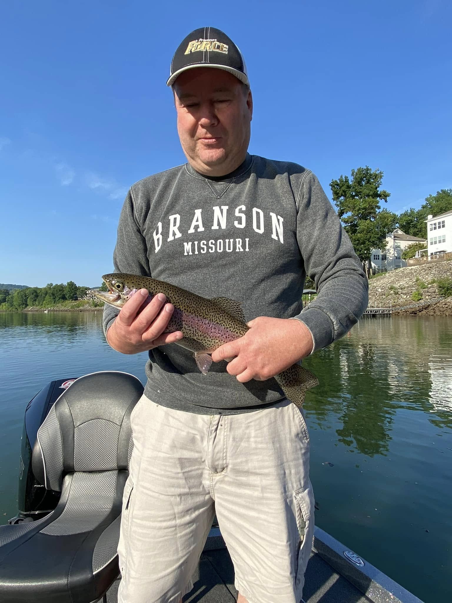 A man holding a small rainbow trout on a boat, wearing a Branson Missouri sweatshirt and a cap, on a clear day by a lake.