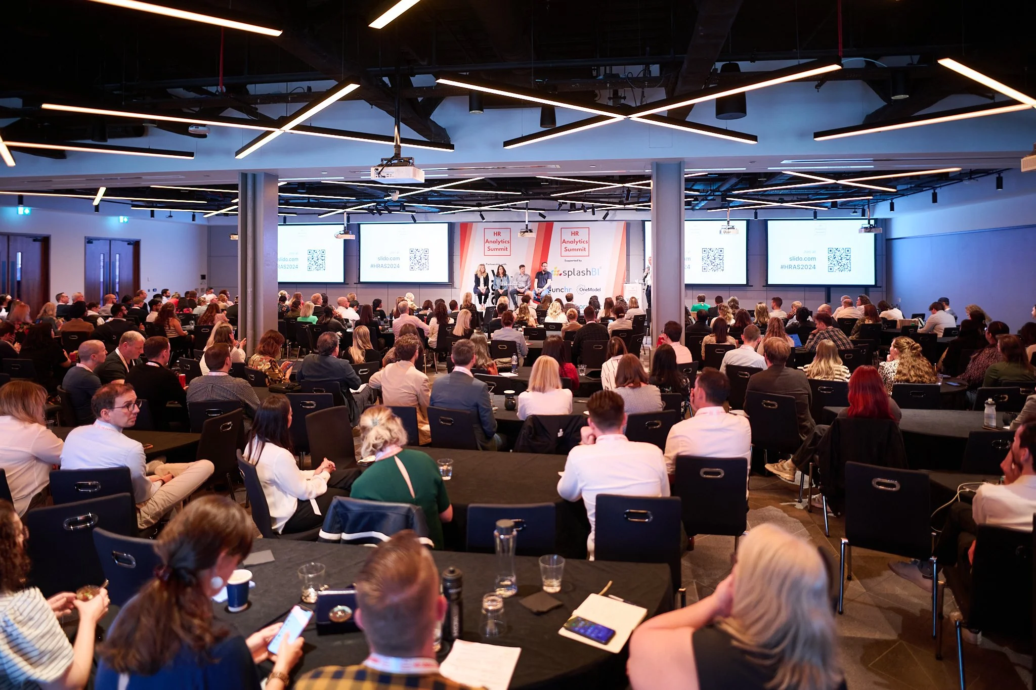 Conference attendees listening to a panel discussion at the HR Analytics Summit 2024, with presenters on stage and screens displaying information.