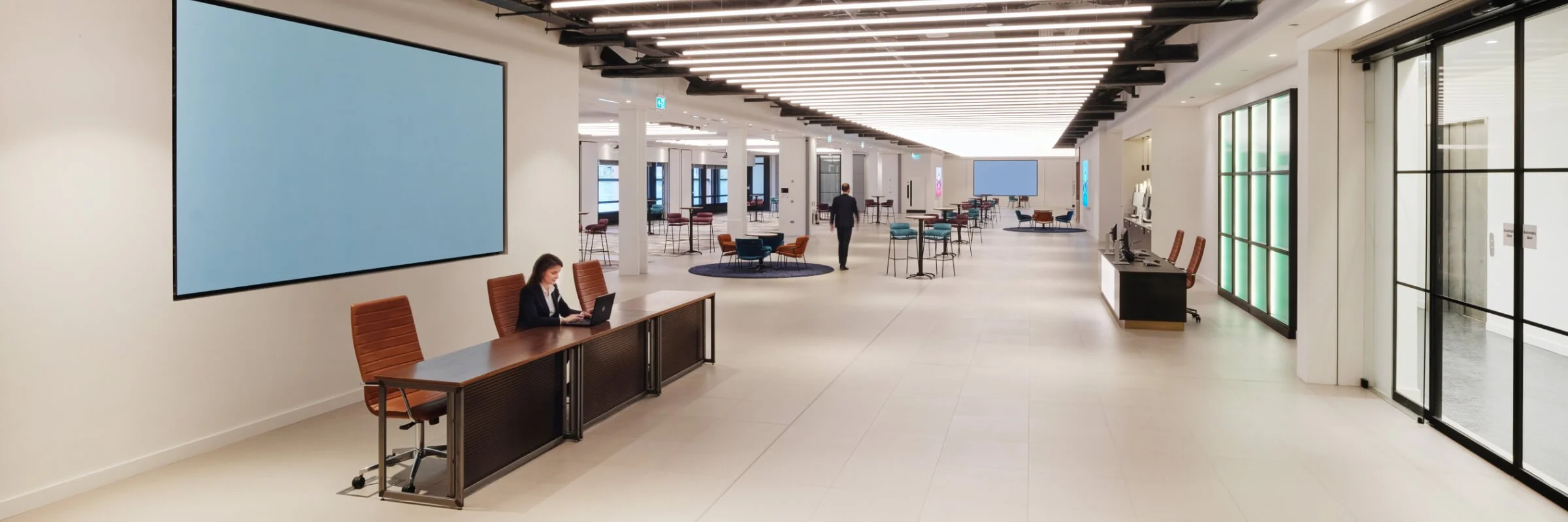 Modern office interior with a long corridor featuring large screens, desks with brown chairs, lounge seating, and overhead lighting. A person is working on a laptop at a desk.