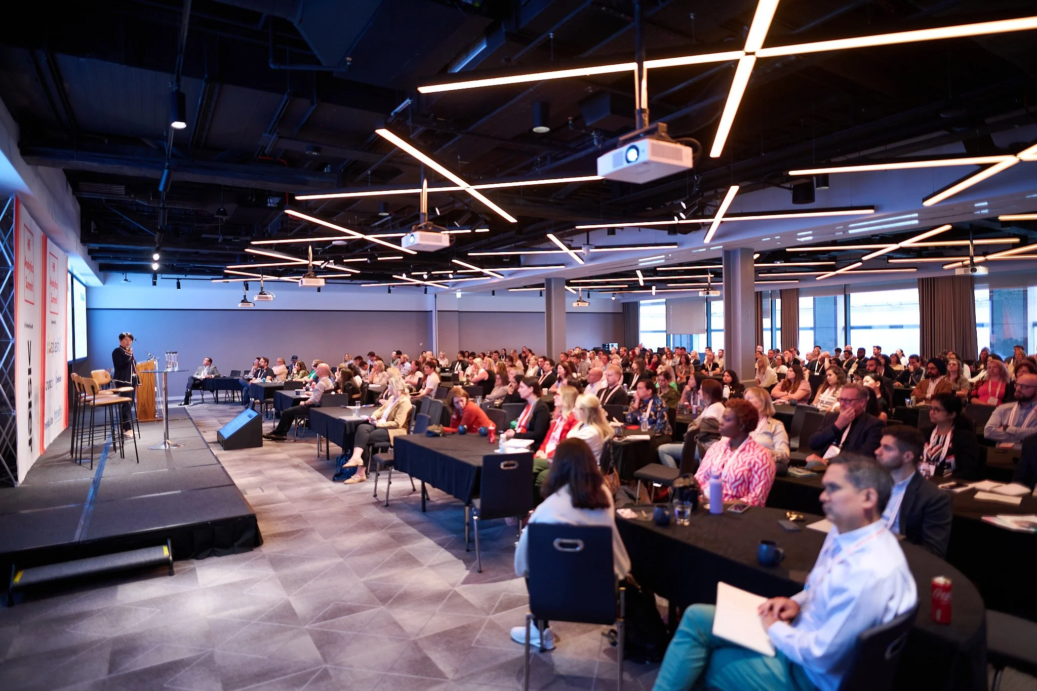 Conference room with a presenter speaking on stage to a seated audience.