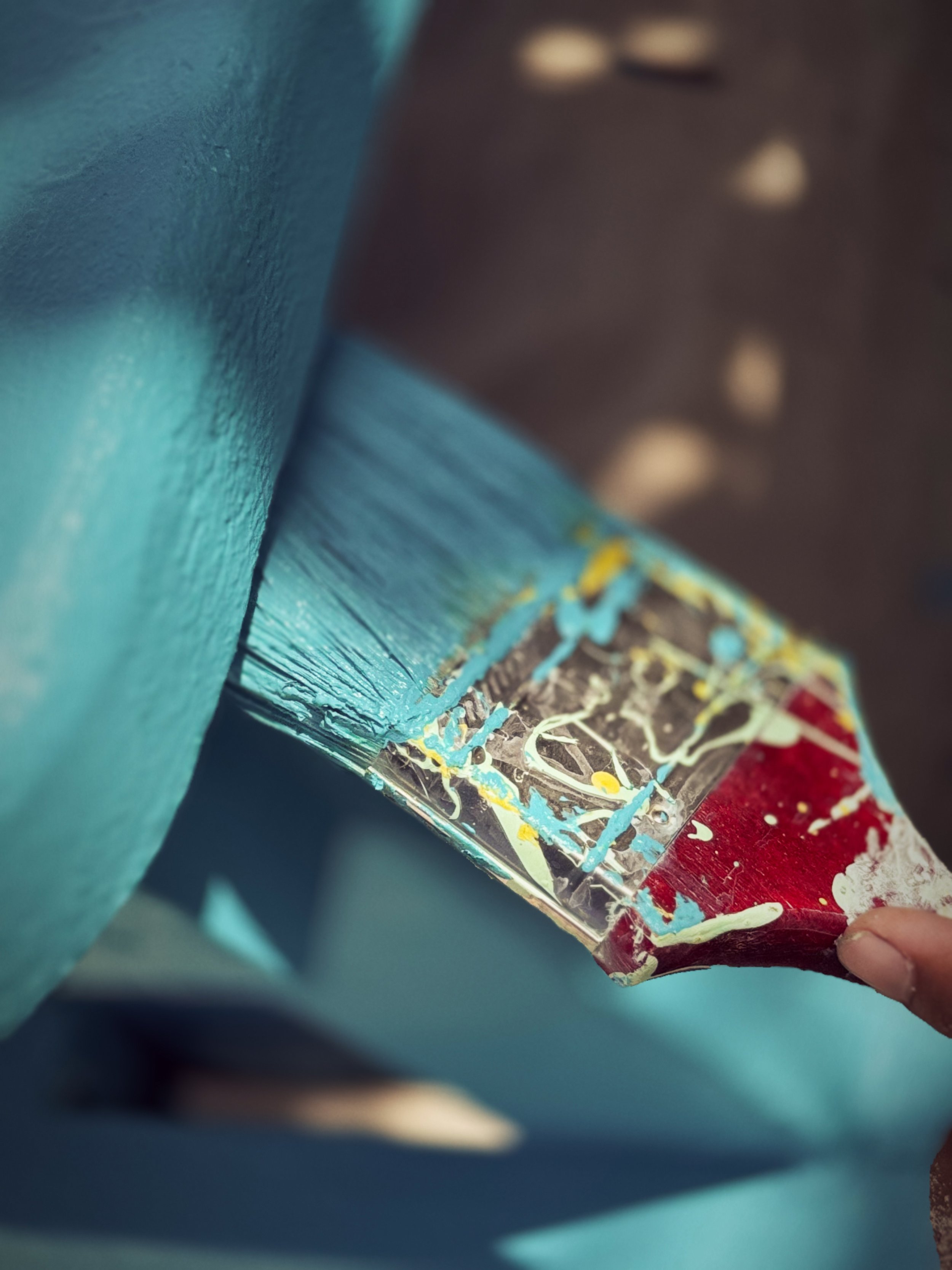 Close-up of a hand holding a paint scraper with blue, yellow, white, and red paint splatters, next to a blue painted surface.