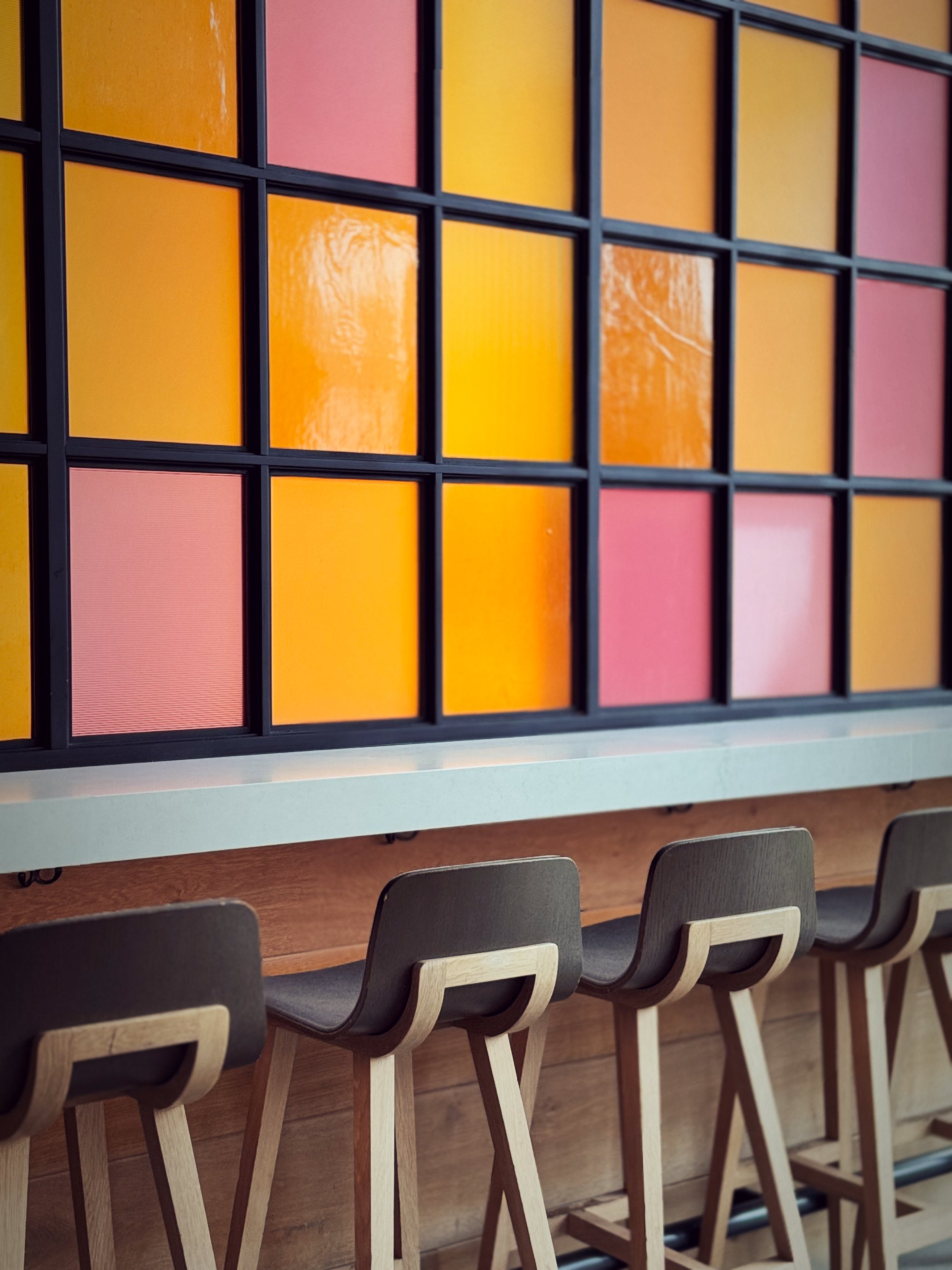 A row of bar stools underneath a wooden counter with a colorful stained glass window featuring yellow and pink panes.